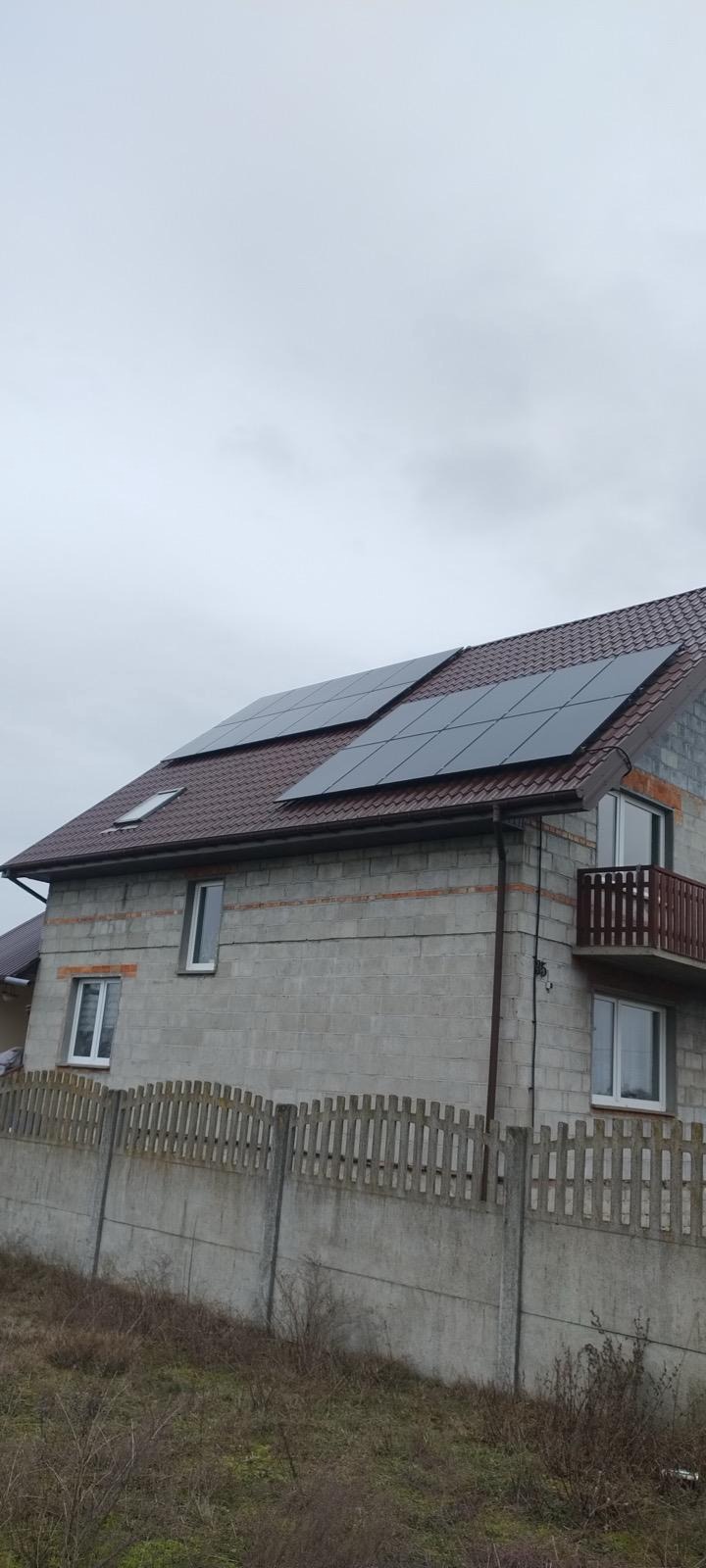 Dom w trakcie budowy z zamontowanymi panelami fotowoltaicznymi na brązowym dachu. Widoczne szare ściany z pustaków i drewniany balkon.