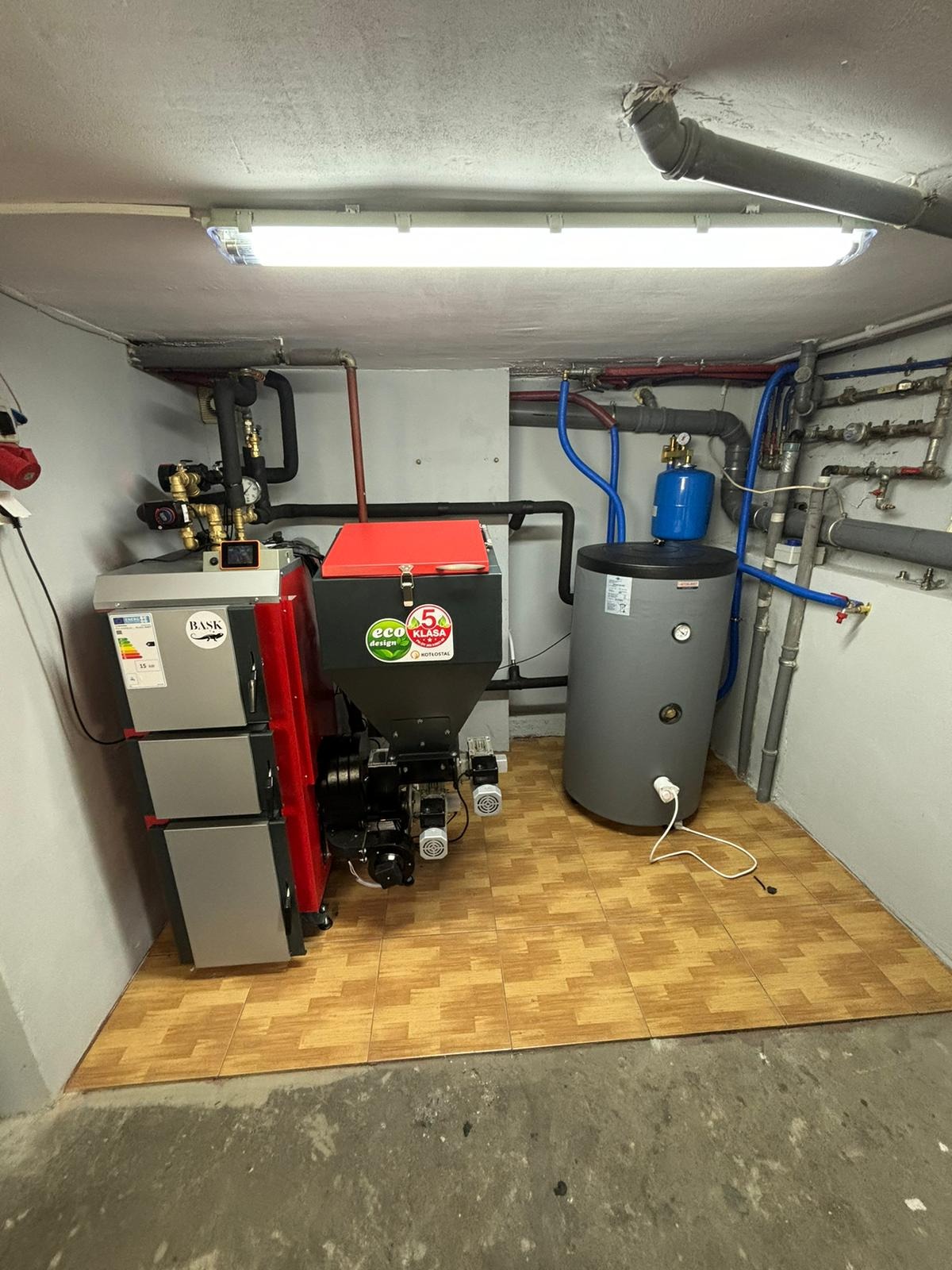 Zmontowany kocioł na ekogroszek z zasobnikiem, zbiornik wyrównawczy i liczne rury w kotłowni z wyłożoną podłogą. Widoczne elementy instalacji grzewczej.