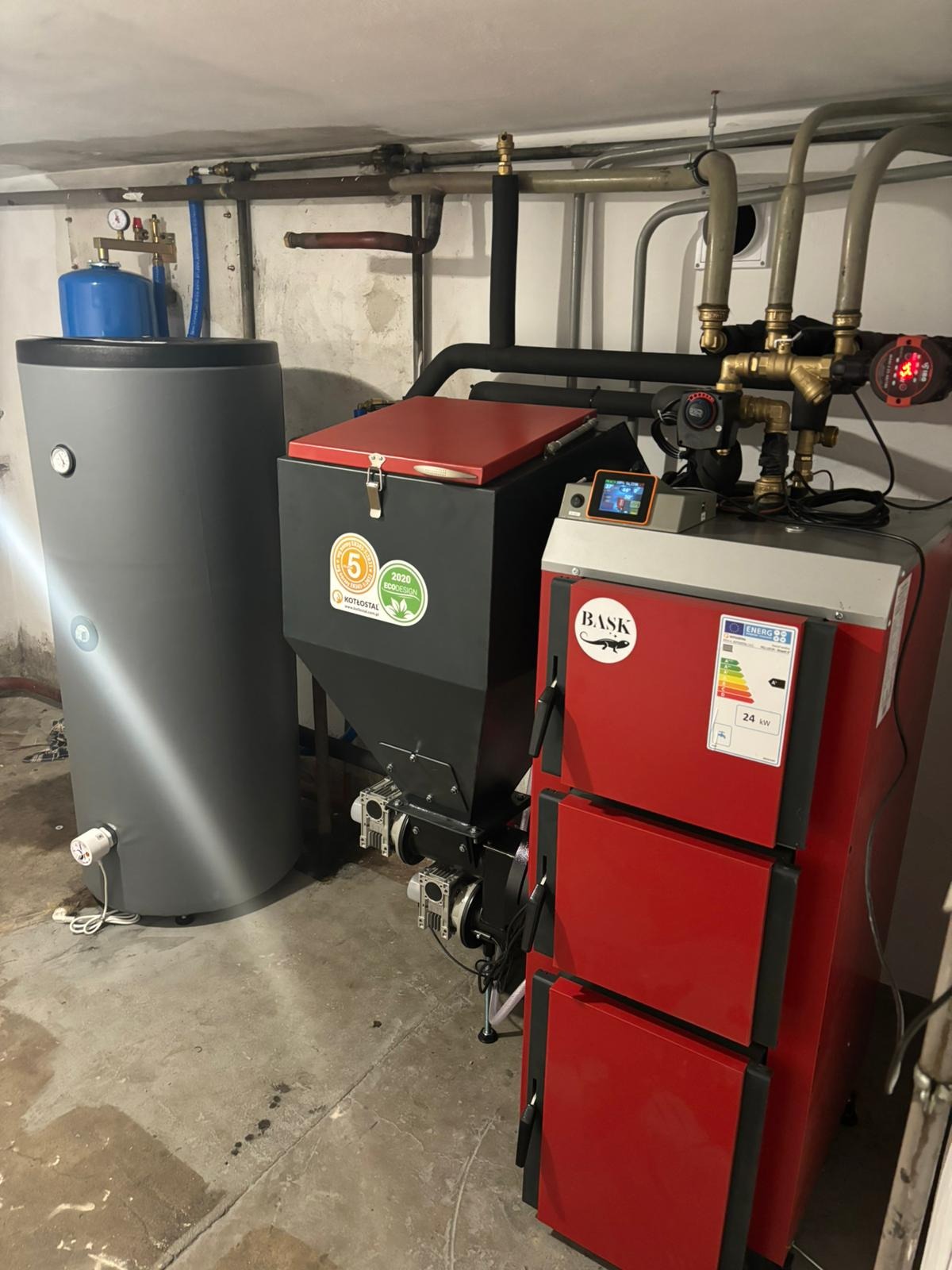 Nowoczesny, zainstalowany piec CO z podajnikiem i zasobnikiem na paliwo stałe, widoczne rury instalacji grzewczej. Kotłownia w piwnicy.