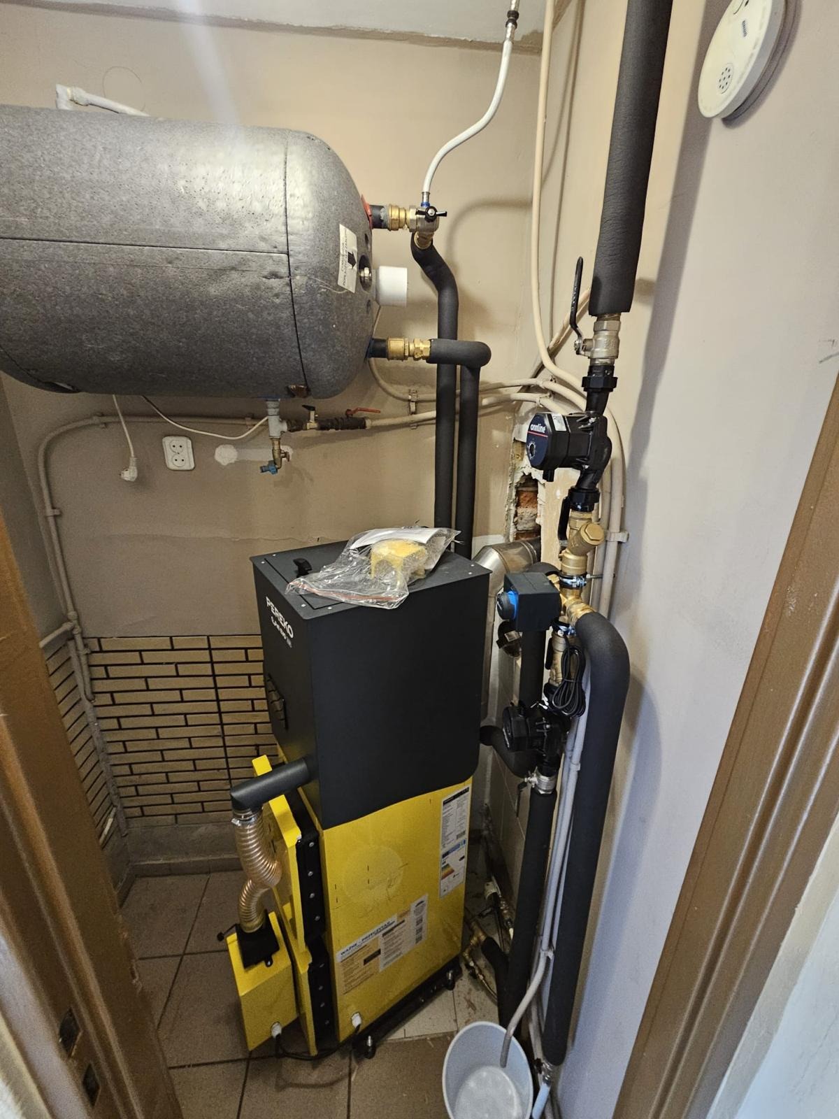 Żółto-czarny piec Pereko KSW Master z podajnikiem, zbiornik wody, rury instalacji grzewczej w kotłowni. Widoczne elementy instalacji i podłączenia.