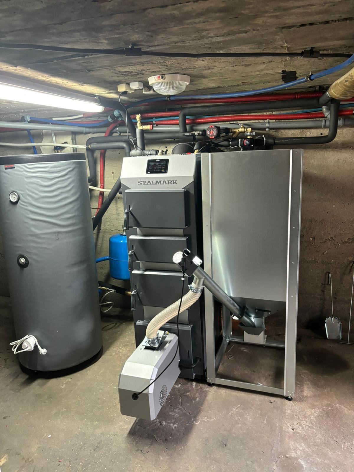 Szary piec Stalmark z podajnikiem i zbiornikiem na wodę w kotłowni. Widoczne rury instalacji grzewczej pod sufitem. Oświetlenie jarzeniowe.