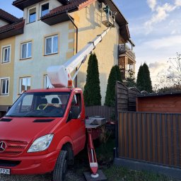KlimaFlow - Czerwony podnośnik koszowy Mercedes przy budynku. Pracownik montuje klimatyzator na elewacji. Widoczne okna, balkon i ogrodzenie. Błękitne niebo z chmurami w tle.