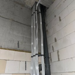 KlimaFlow - Montaż instalacji rekuperacji w surowym wnętrzu: białe rury wentylacyjne przymocowane do ściany z betonu komórkowego, szara rura kanalizacyjna. Widok z dołu.