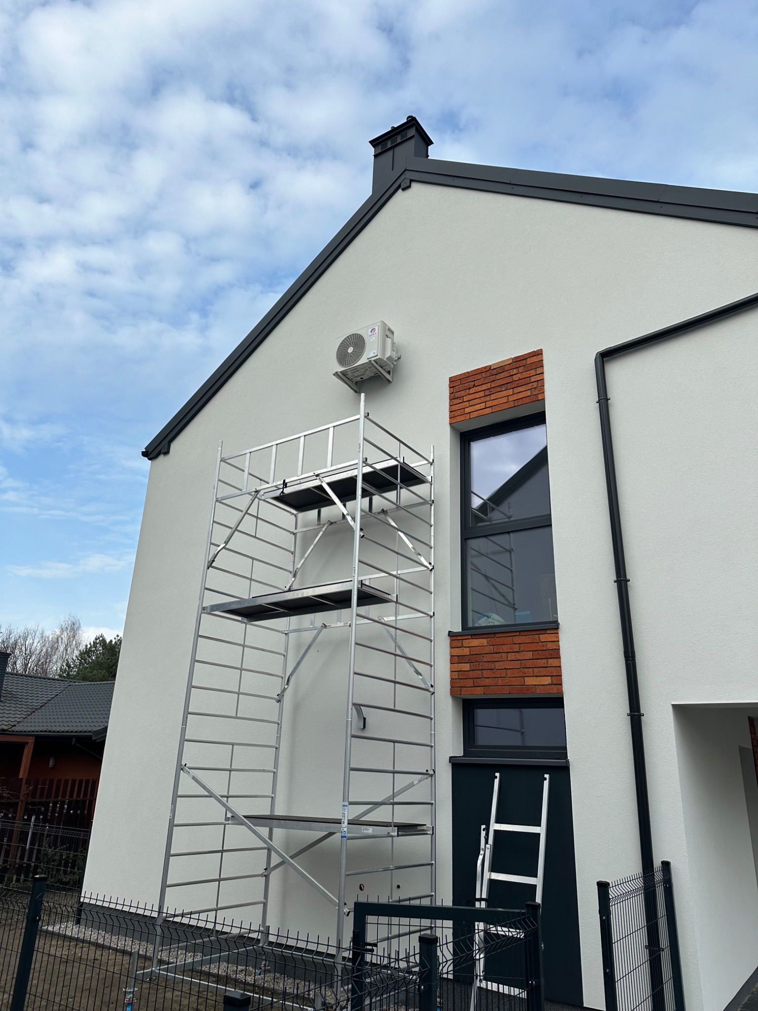 Biały budynek z zamontowaną jednostką zewnętrzną klimatyzacji, widoczne rusztowanie i drabina, sugerujące trwający montaż. Ujęcie z dołu, błękitne niebo w tle.