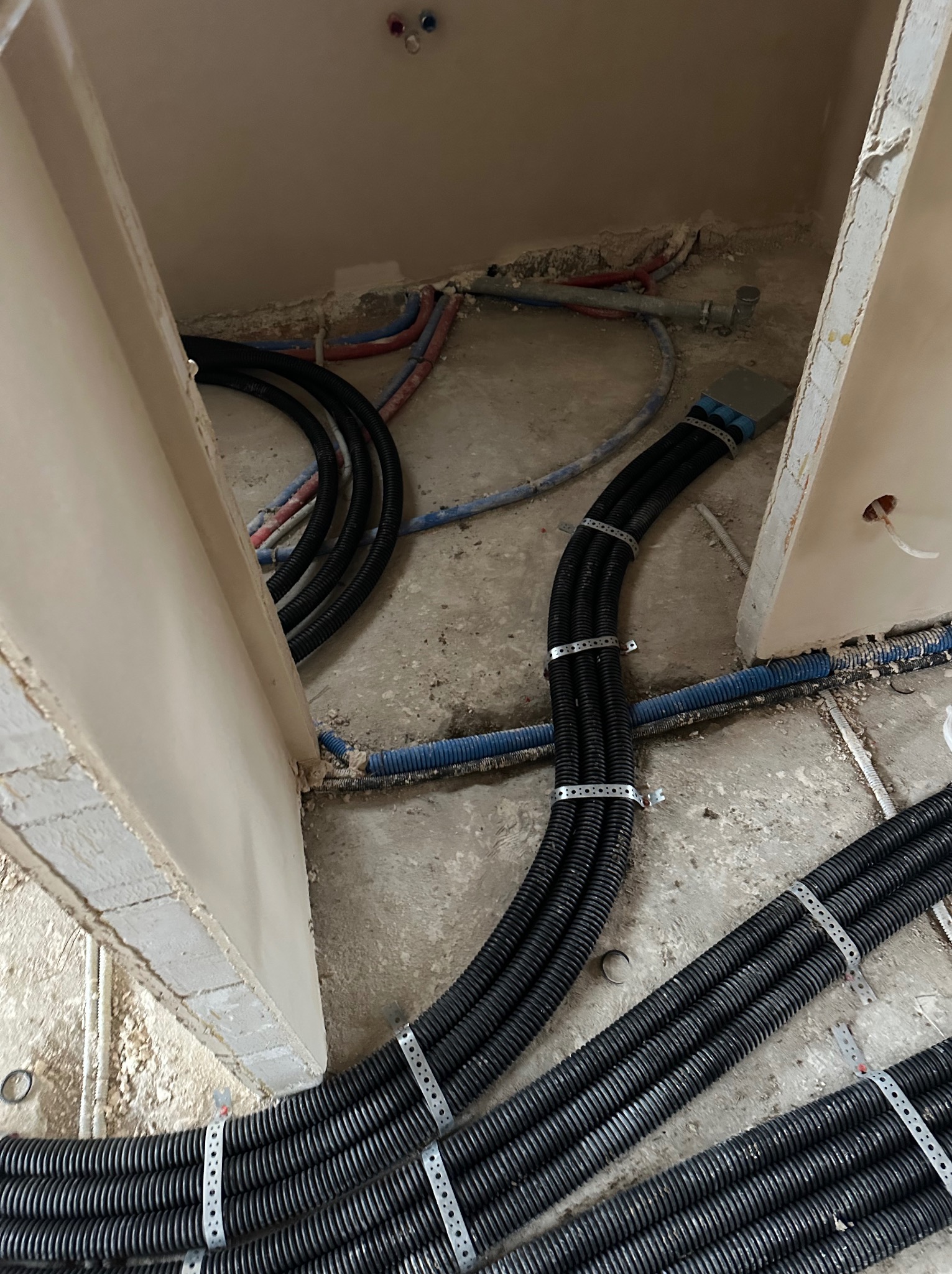 Układanie czarnych, karbowanych rur wentylacyjnych na podłodze w trakcie montażu. Widoczne elementy mocujące i inne instalacje w tle. Surowy stan wykończenia.