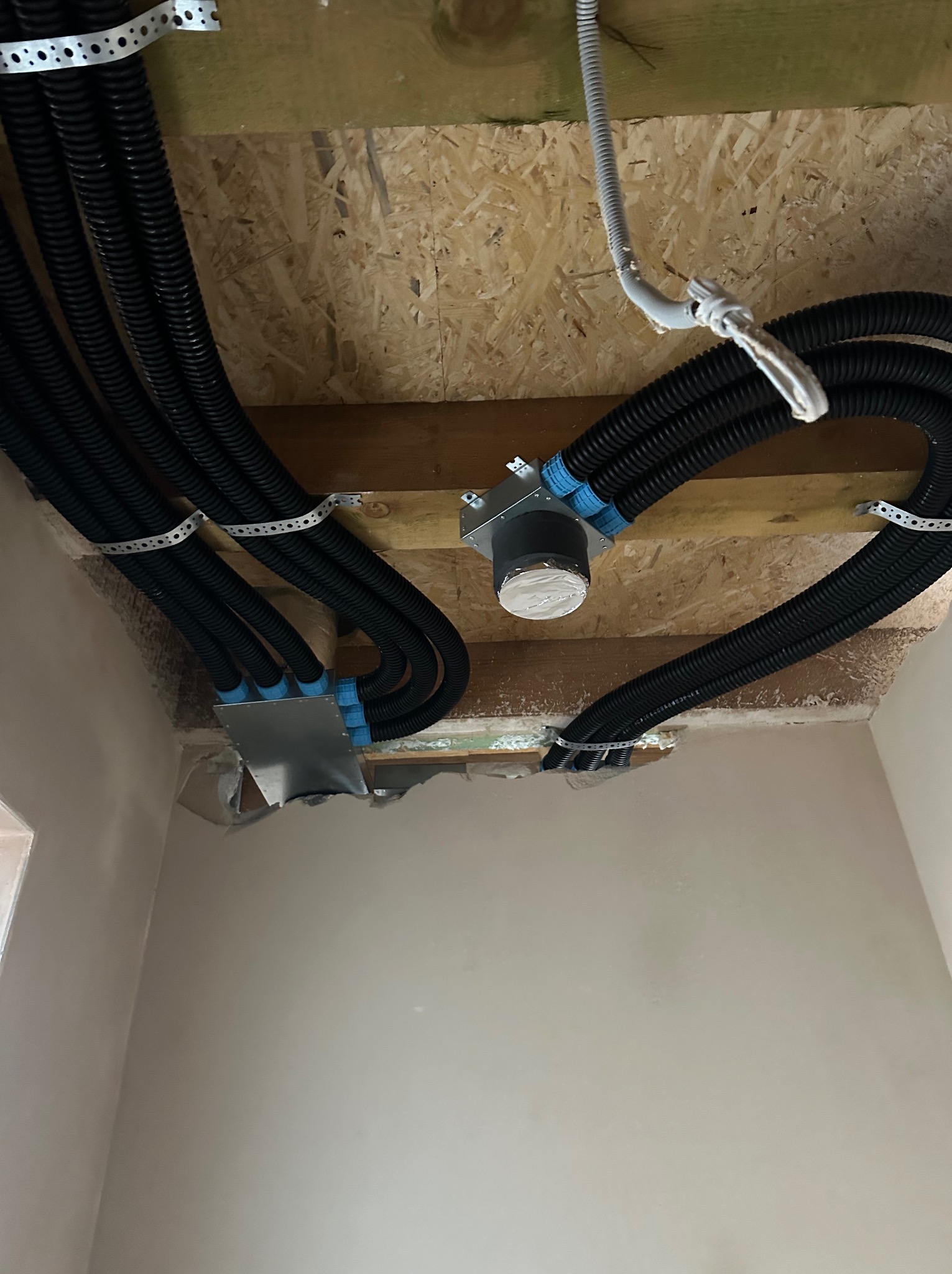 Czarny system rur wentylacyjnych zamontowany pod drewnianym stropem, widoczne elementy mocujące i skrzynki rozdzielcze. Instalacja w trakcie prac wykończeniowych.