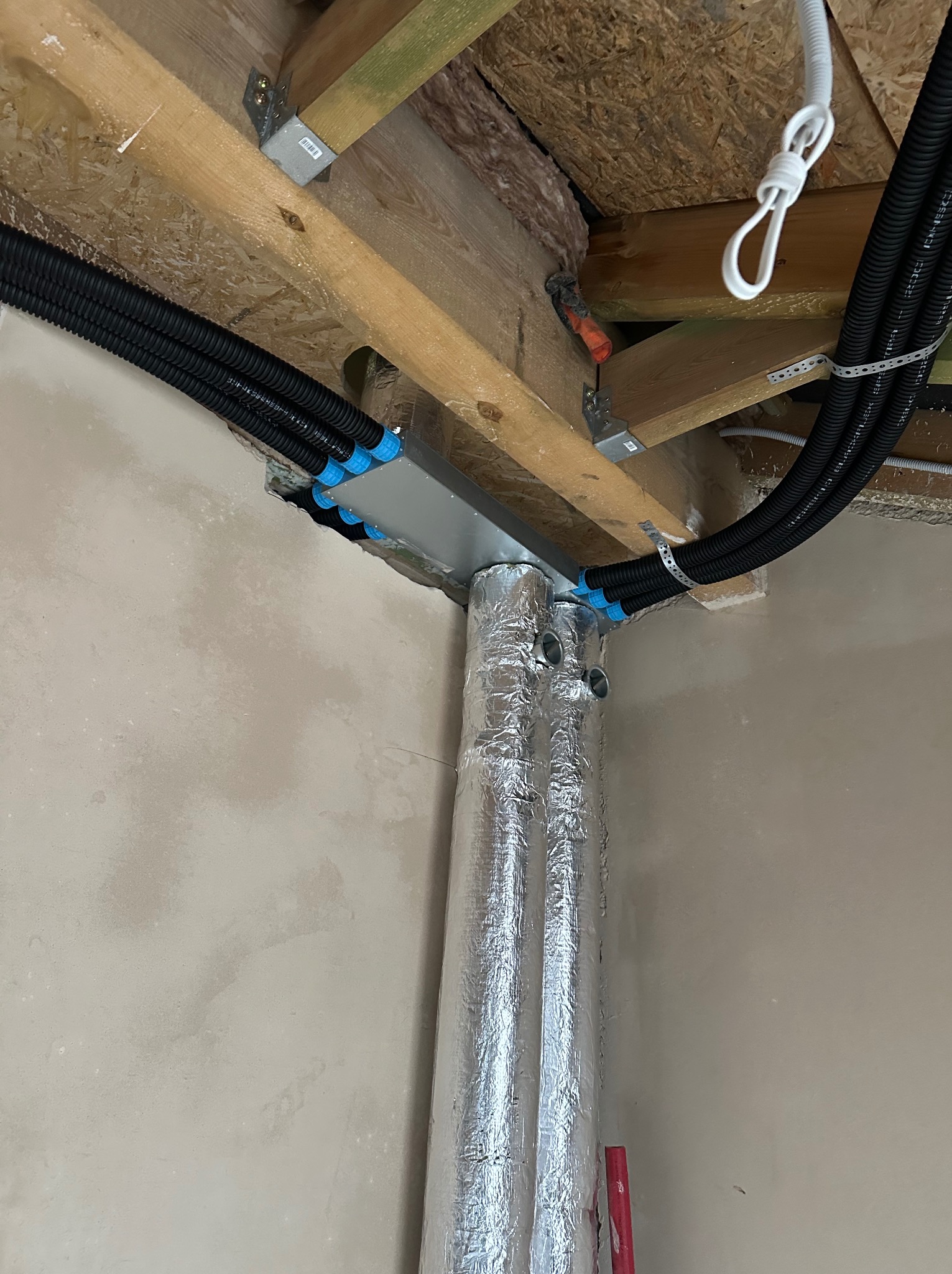 Szczegółowy widok instalacji rekuperacji z czarnymi rurami i srebrną izolacją, zamontowanej pod drewnianym stropem w surowym wnętrzu.