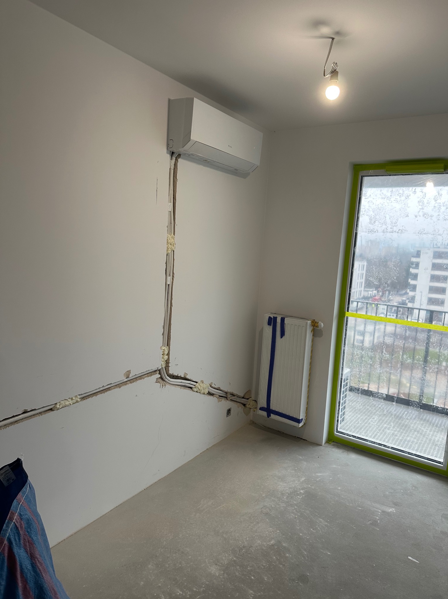 Biały klimatyzator na ścianie w surowym wnętrzu. Widoczne instalacje, rury zabezpieczone pianką montażową. Okno z widokiem na budynek. Grzejnik zabezpieczony taśmą.
