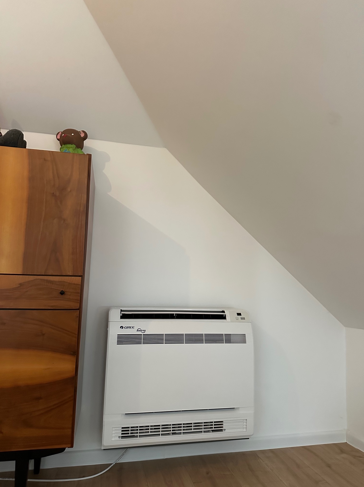 Biały klimatyzator podłogowy marki Gree, zamontowany w rogu pokoju na poddaszu, obok drewnianej komody. Widoczny kabel zasilający. Na komodzie dekoracje.