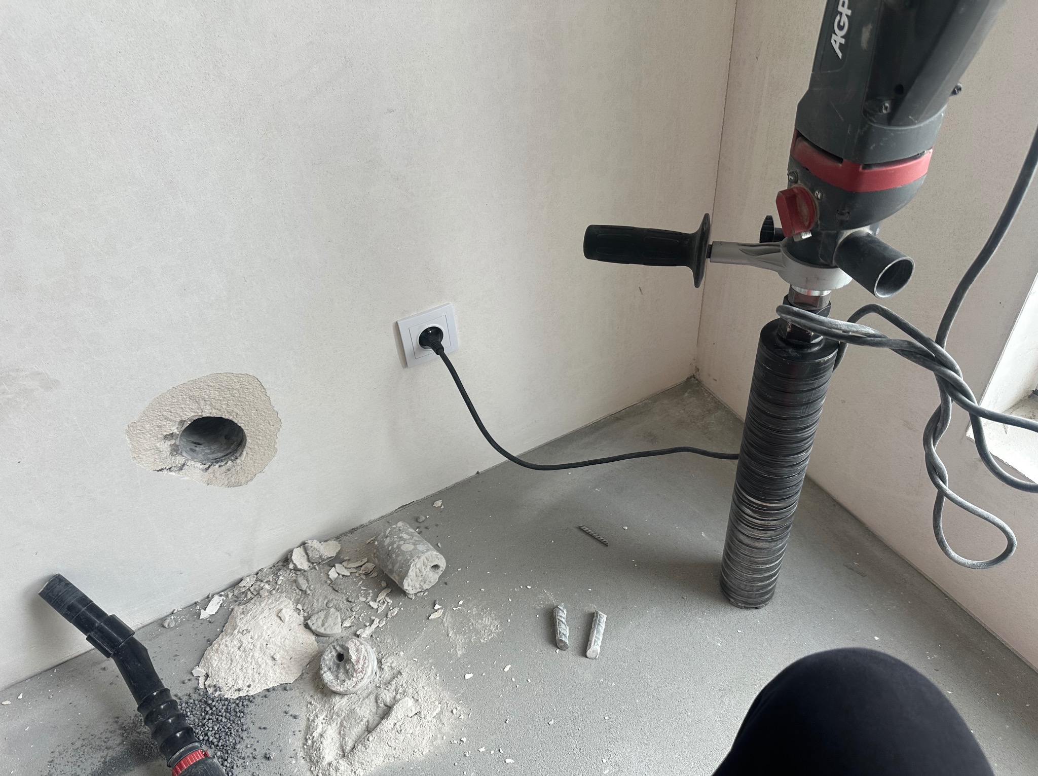 Wiercenie otworu w ścianie pod instalację wentylacyjną za pomocą wiertnicy diamentowej. Widoczne narzędzia, otwór w ścianie i resztki materiału.