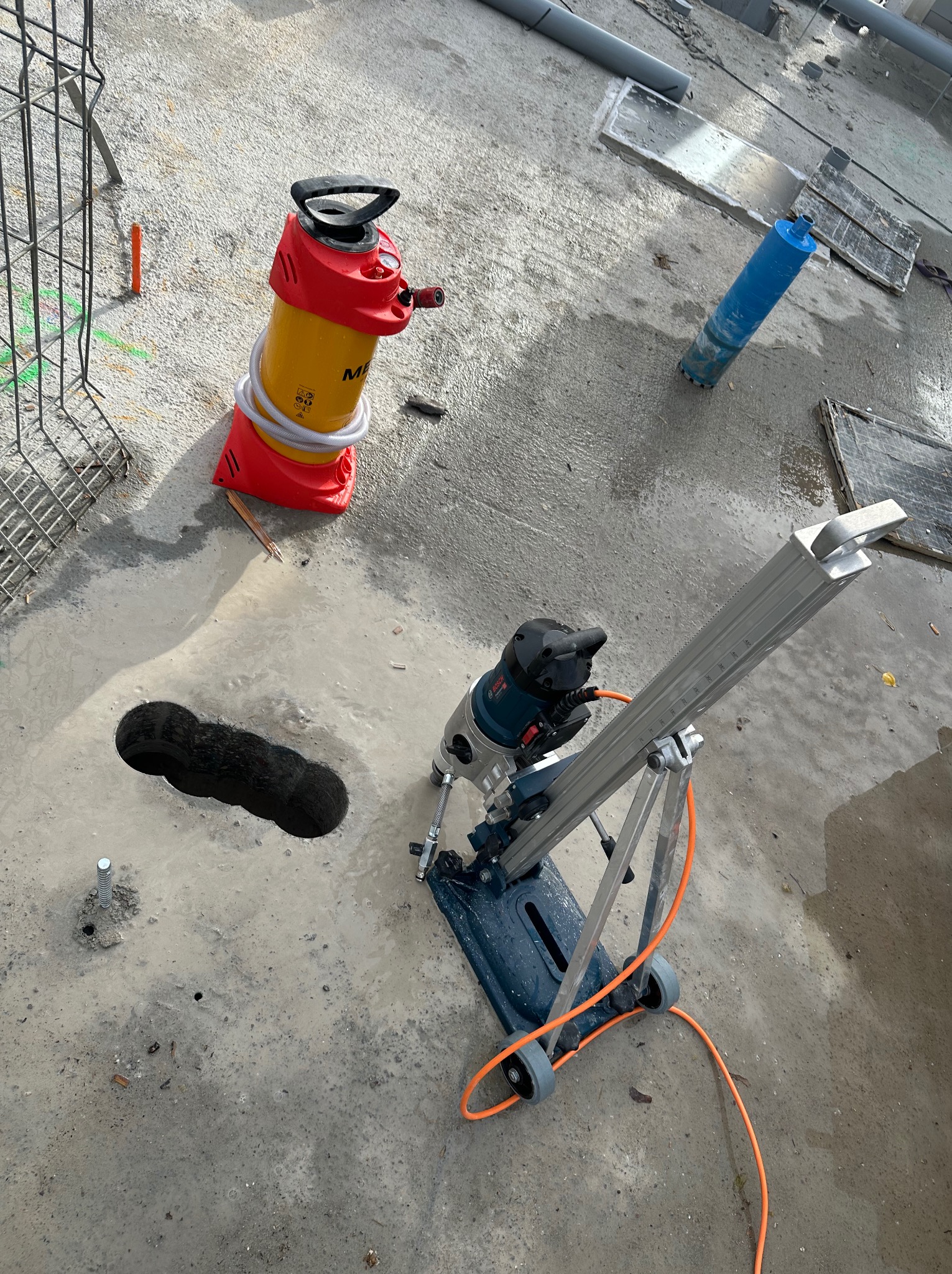 Wiercenie otworów w betonie wiertnicą diamentową z odsysaniem, widoczny rdzeń. Praca na mokro, żółto-czerwony odkurzacz przemysłowy obok. Szara, betonowa powierzchnia.