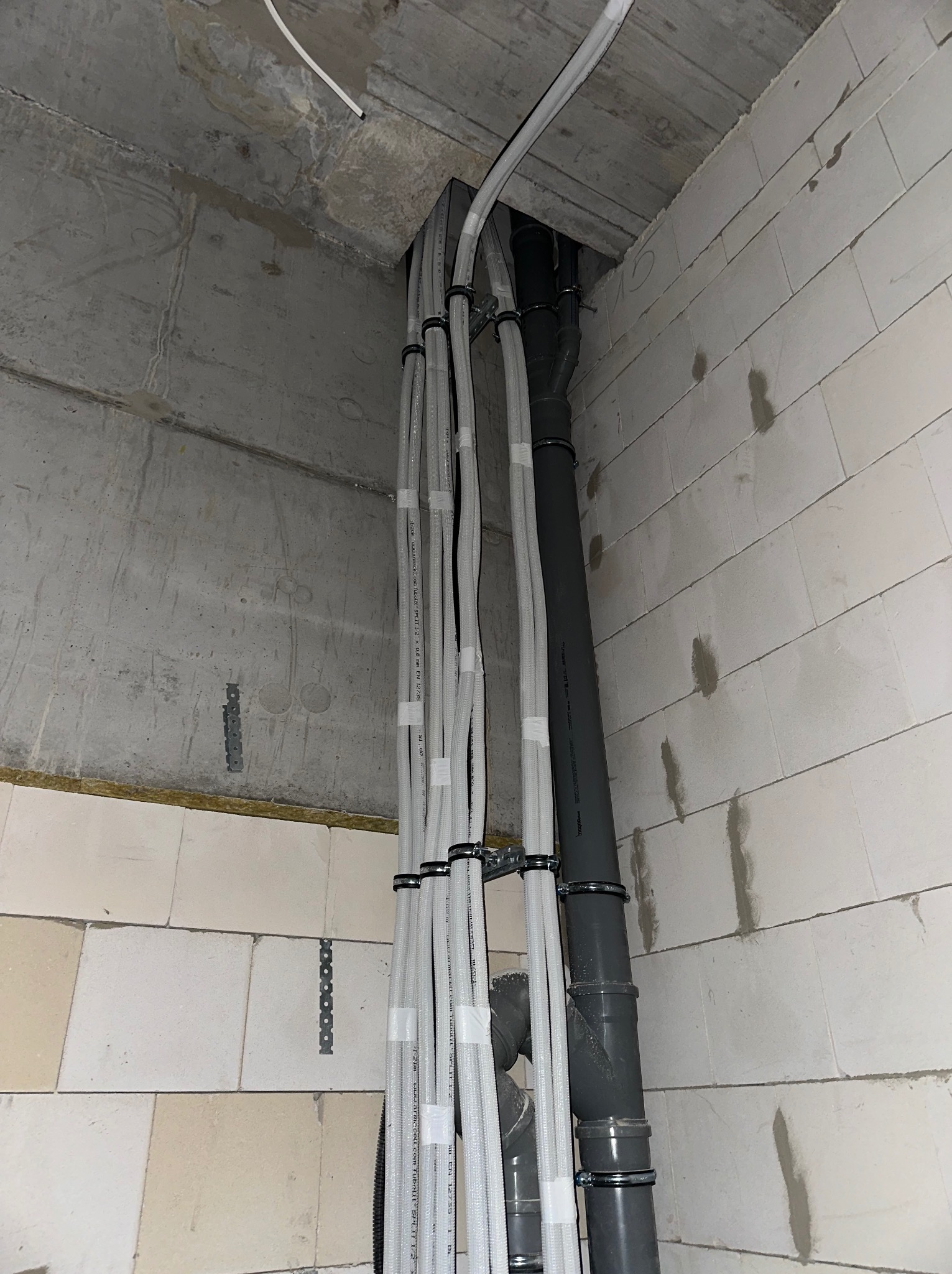 Montaż instalacji rekuperacji w surowym wnętrzu: białe rury wentylacyjne przymocowane do ściany z betonu komórkowego, szara rura kanalizacyjna. Widok z dołu.