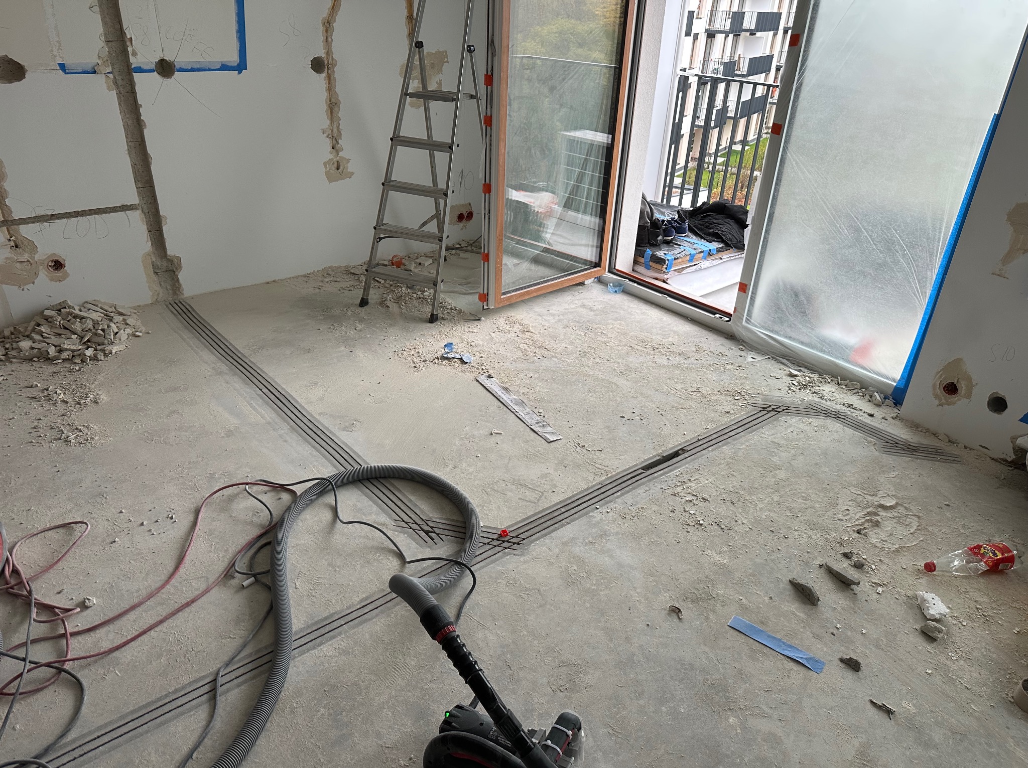 Przygotowanie podłoża pod instalację wentylacyjną: wycięte bruzdy w posadzce, widoczny odkurzacz przemysłowy i drabina. Balkon z widokiem na budynek w tle.