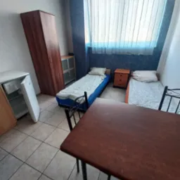 Pokój do wynajęcia z dwoma łóżkami, szafą, stolikiem i lodówką. Widok na okno z żaluzjami pionowymi. Całość w neutralnej kolorystyce.