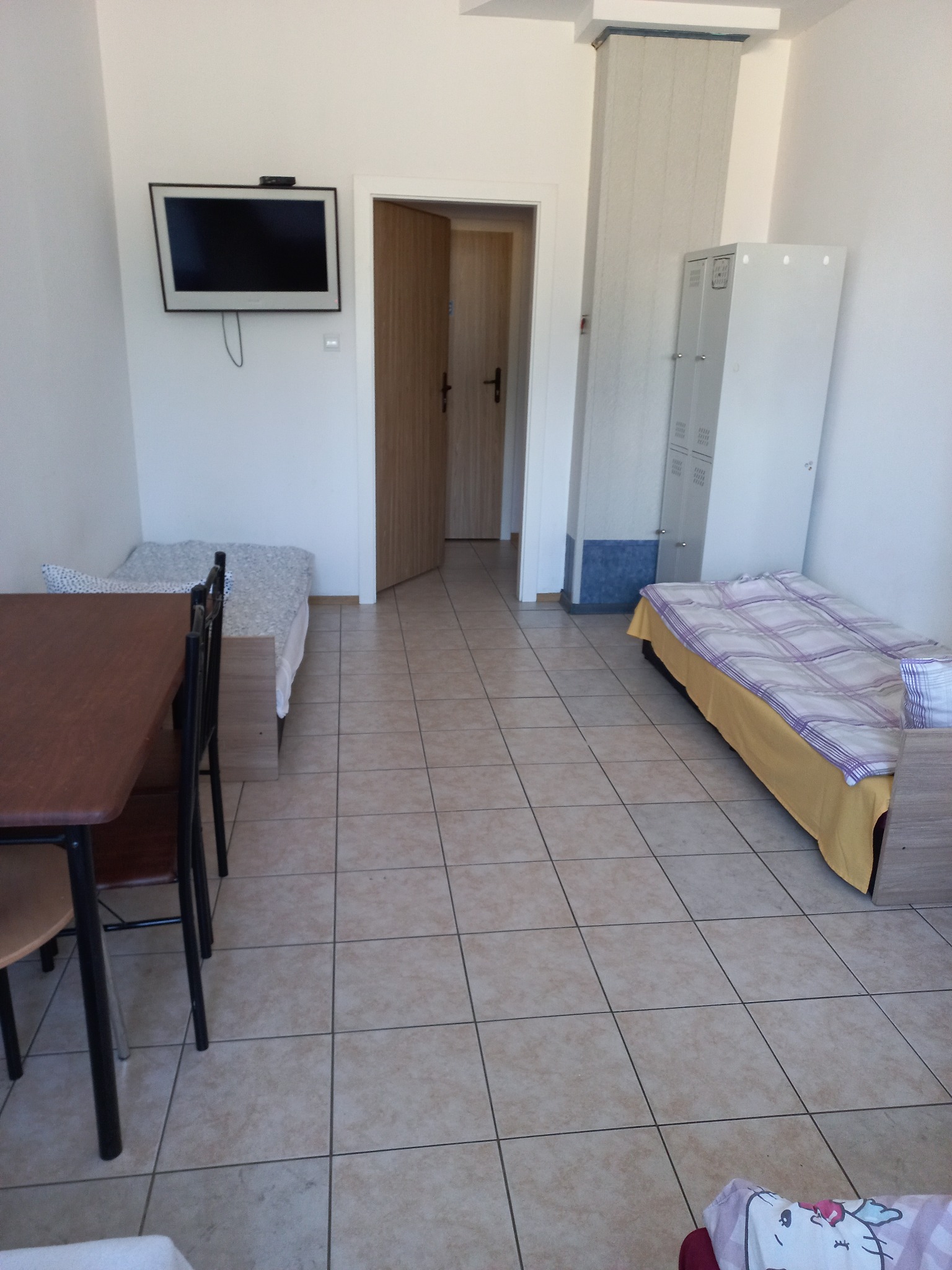 Pokój z dwoma łóżkami, stołem i telewizorem na ścianie. Widoczne szafki i drzwi do kolejnego pomieszczenia. Jasne wnętrze, płytki na podłodze.