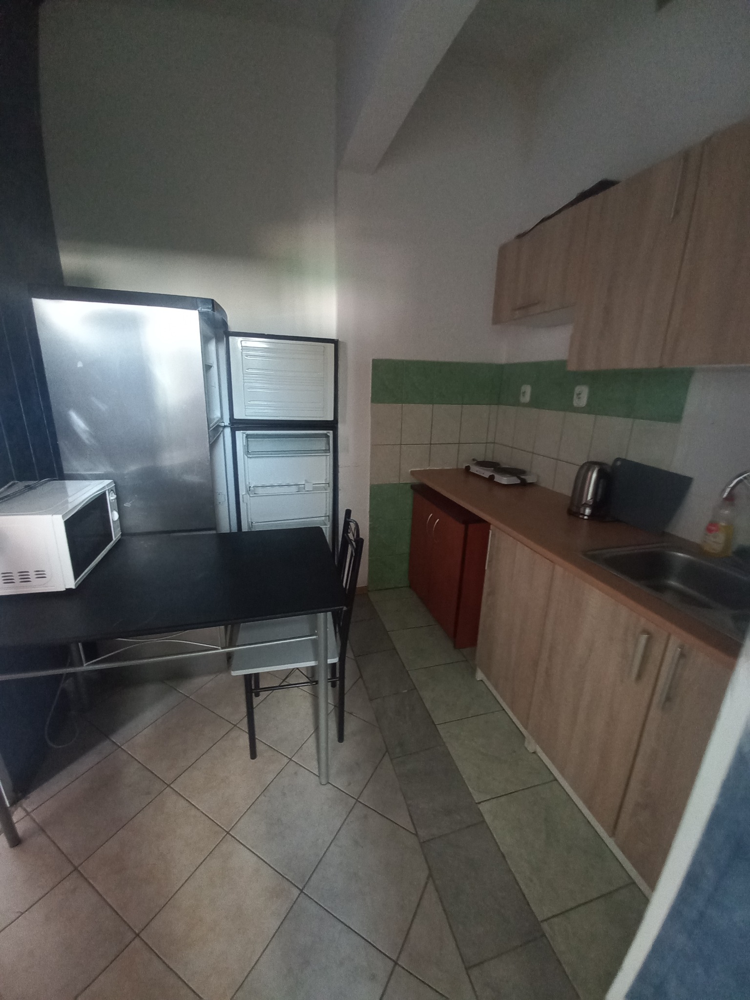 Kuchnia w wynajmowanym studio: lodówka, mikrofalówka, stół z krzesłami, szafki kuchenne w odcieniach drewna, zielono-białe płytki na ścianie, płytki podłogowe.