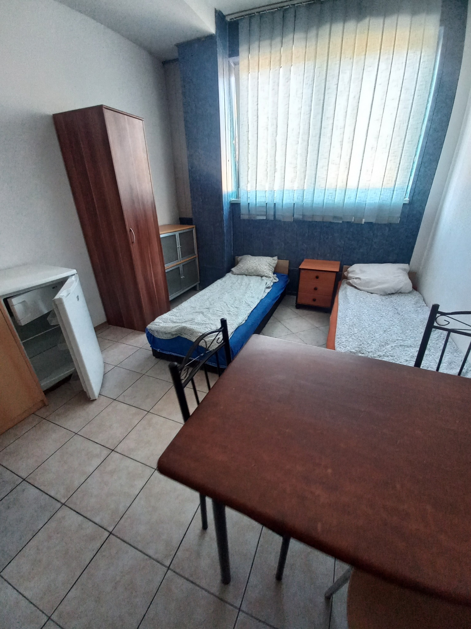 Pokój do wynajęcia z dwoma łóżkami, szafą, stolikiem i lodówką. Widok na okno z żaluzjami pionowymi. Całość w neutralnej kolorystyce.