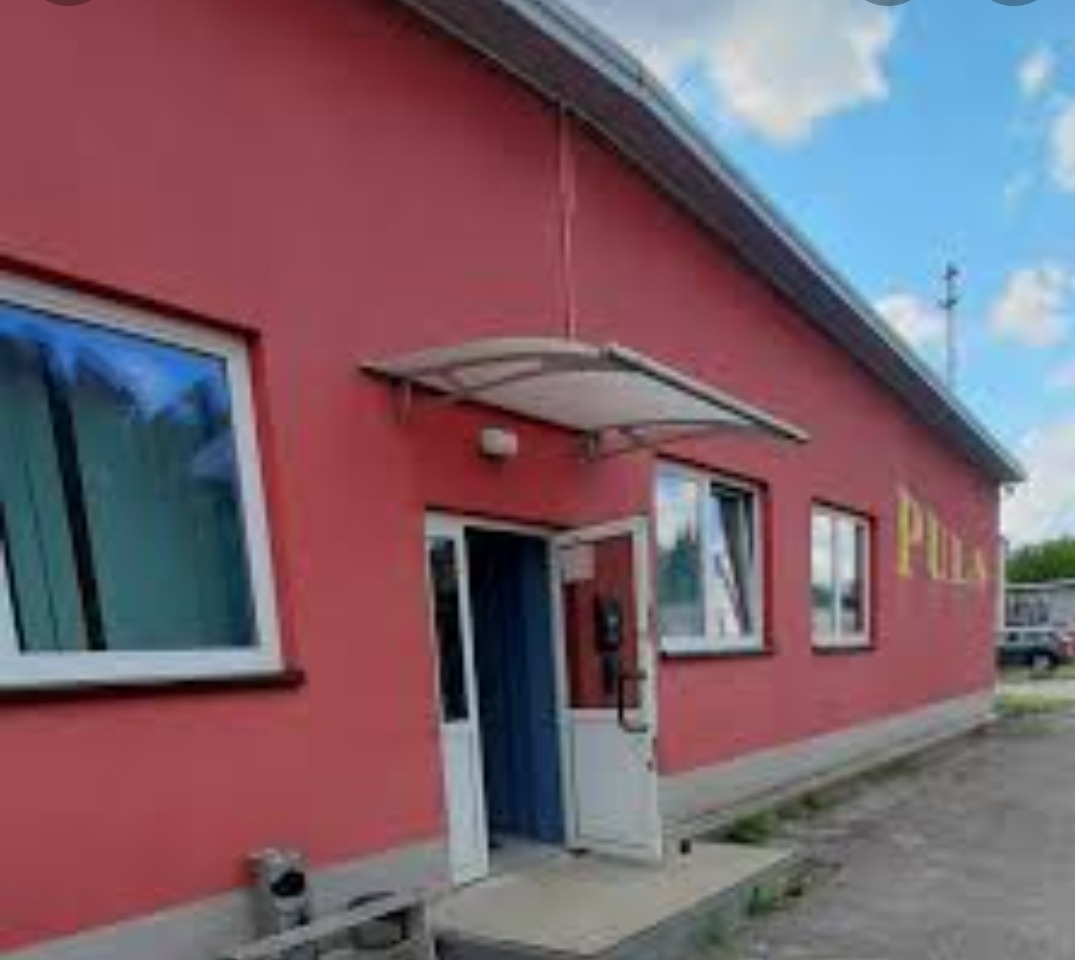 Czerwony budynek z białymi oknami i daszkiem nad wejściem. Widoczny napis 'PULA' na ścianie. Obok parkingu stoi samochód.