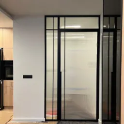 Nowoczesne, rozwierane drzwi ze szkła ornamentowego w czarnej, stalowej ramie. Widoczne zabezpieczenia podłogi na czas montażu. Minimalistyczne wnętrze.