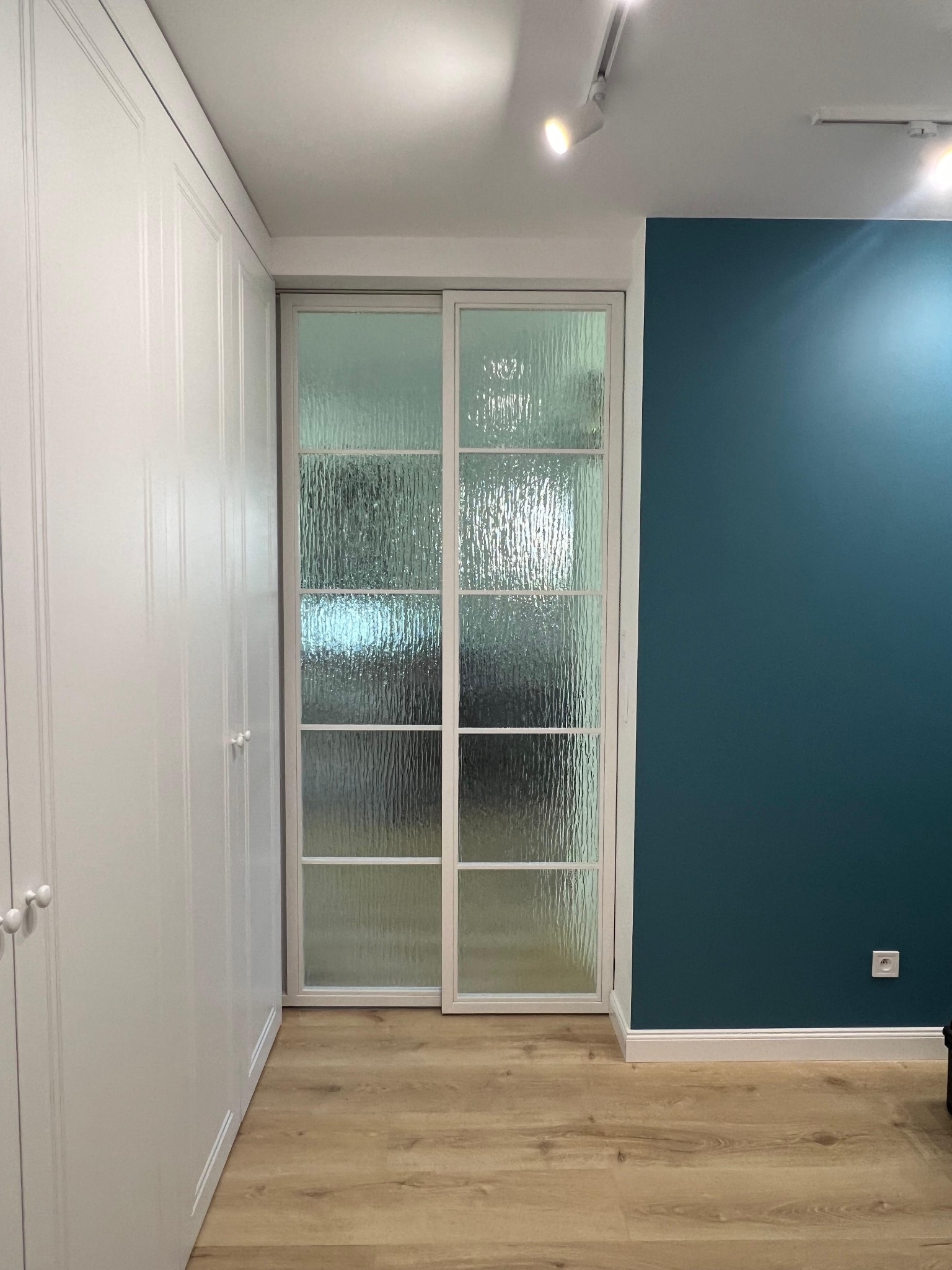 Przesuwne drzwi z ornamentowym szkłem w białej ramie, wkomponowane między białą szafę a ścianę w kolorze ciemnego turkusu. Widoczna drewniana podłoga i oświetlenie.