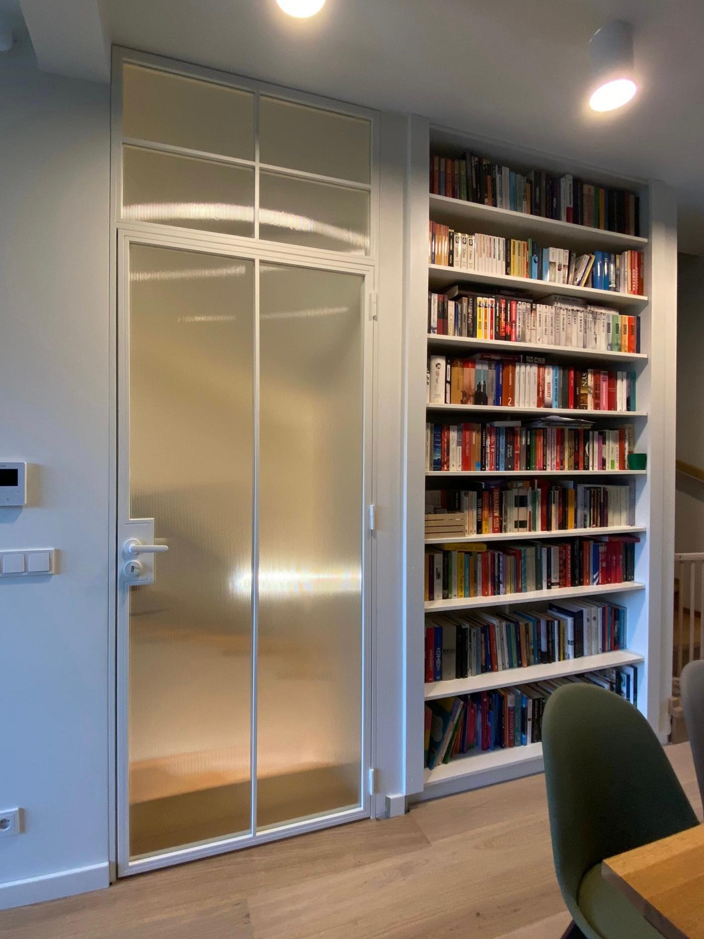 Białe drzwi rozwierane ze szkłem ornamentowym w stalowej ramie, wkomponowane w regał z książkami, oświetlone ciepłym światłem.
