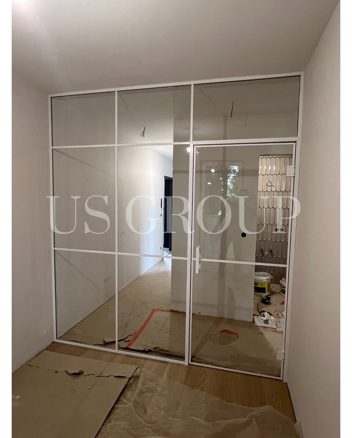 Szklane drzwi rozwierane w białej, stalowej ramie z podziałami na kwadraty. Widoczne zabezpieczenia podłogi podczas montażu.