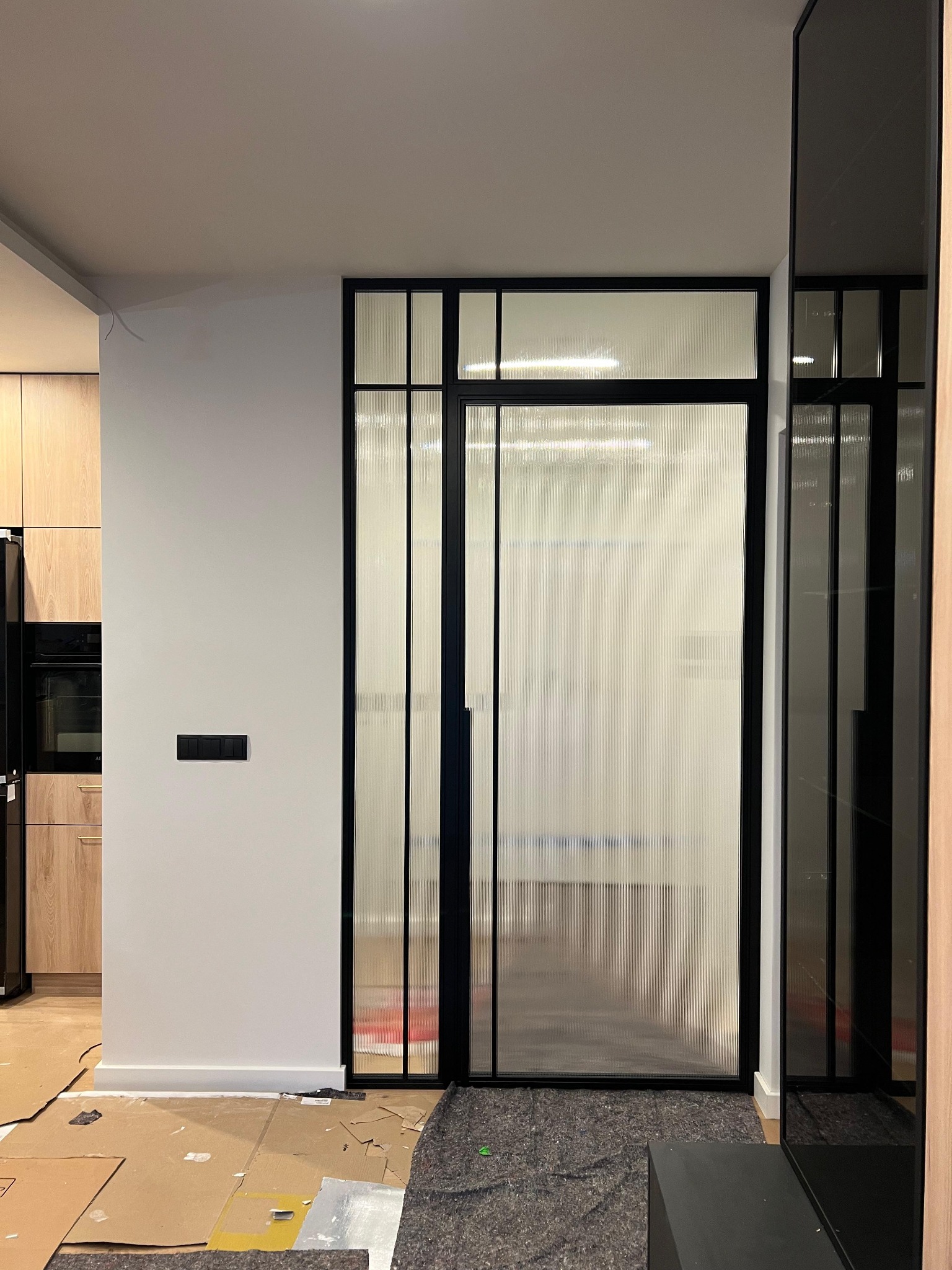 Nowoczesne, rozwierane drzwi ze szkła ornamentowego w czarnej, stalowej ramie. Widoczne zabezpieczenia podłogi na czas montażu. Minimalistyczne wnętrze.