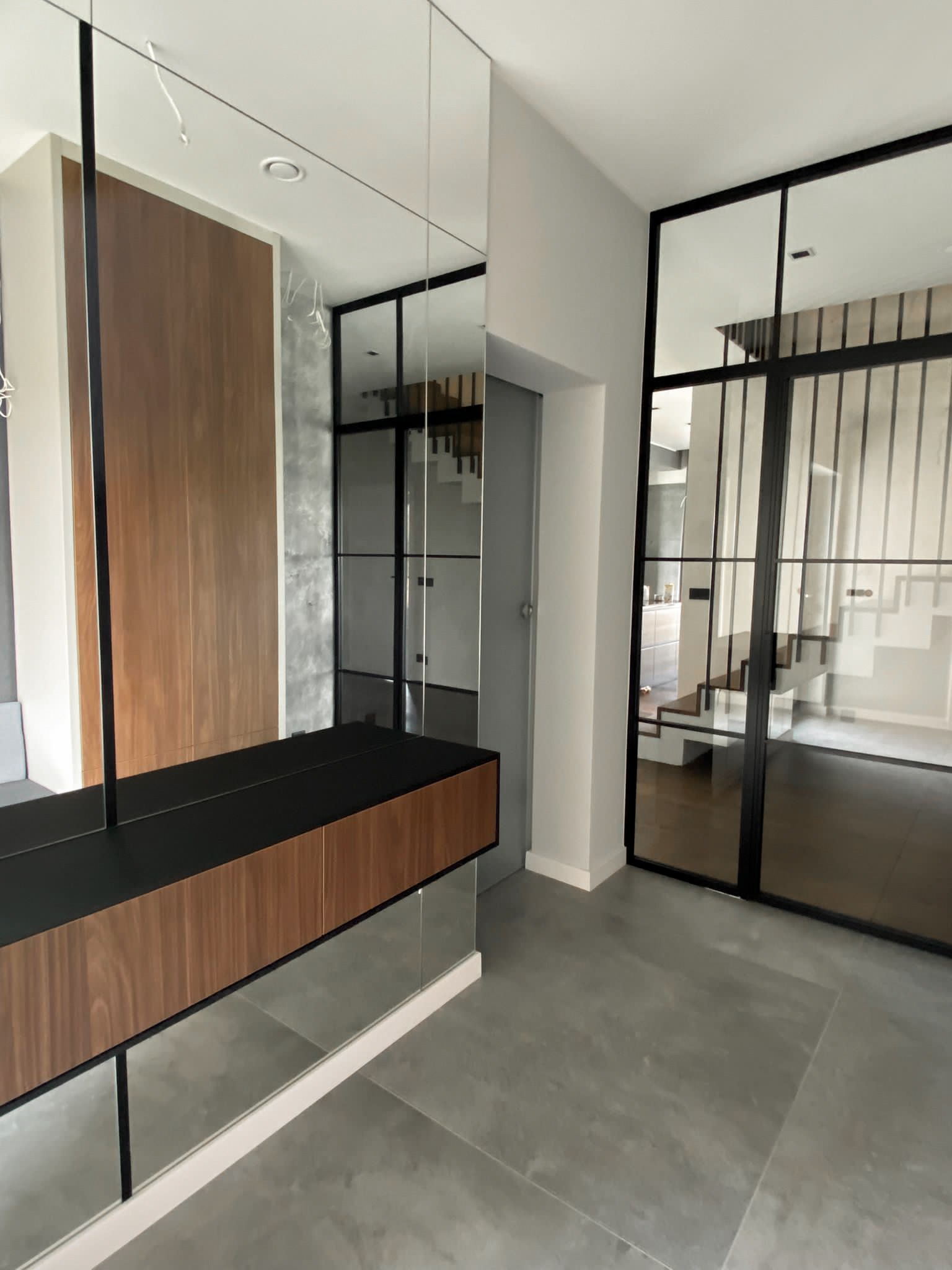 Nowoczesne wnętrze z szklanymi drzwiami w czarnej ramie, widoczne schody i minimalistyczna zabudowa. Transparentne szkło wpuszcza światło do pomieszczenia.