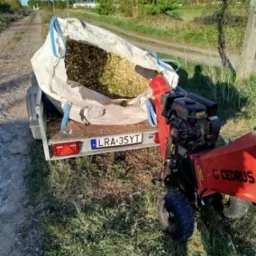 Przyczepa z workiem wypełnionym zrębkami drzewnymi, obok stoi rozdrabniacz do gałęzi marki Cedrus. Widok na polną drogę i łąki w tle.