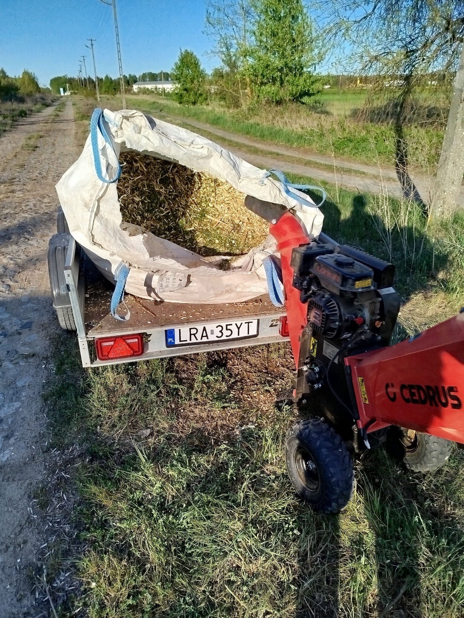 Przyczepa z workiem wypełnionym zrębkami drzewnymi, obok stoi rozdrabniacz do gałęzi marki Cedrus. Widok na polną drogę i łąki w tle.