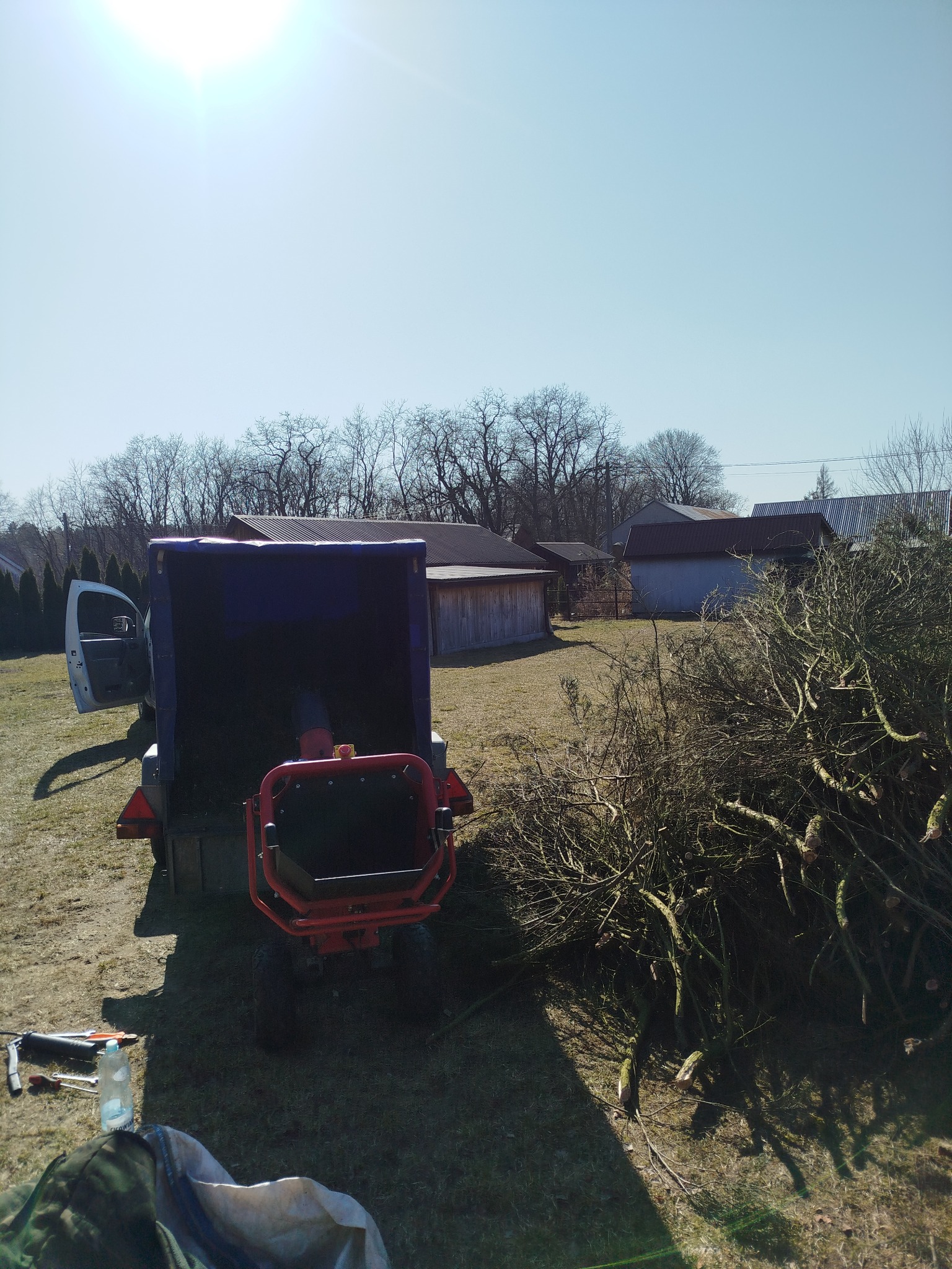 Rozdrabniacz gałęzi z przyczepą wypełnioną pociętymi gałęziami na tle zabudowań gospodarczych w słoneczny dzień. Widoczne narzędzia i pozostałości po cięciu.