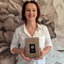 Uśmiechnięta kobieta prezentuje nagrodę/wyróżnienie z 2023 roku, trzymając ją na tle ściany z motywem kwiatowym. Widoczny tekst na nagrodzie.