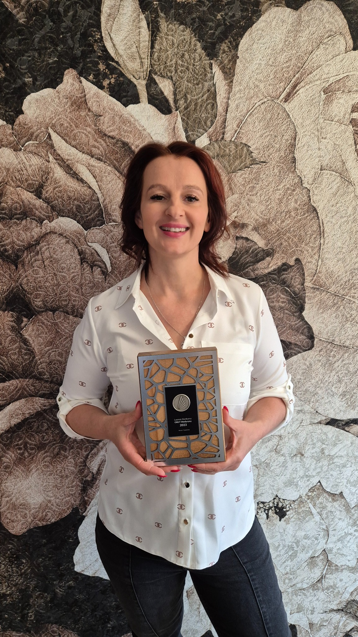 Uśmiechnięta kobieta prezentuje nagrodę/wyróżnienie z 2023 roku, trzymając ją na tle ściany z motywem kwiatowym. Widoczny tekst na nagrodzie.