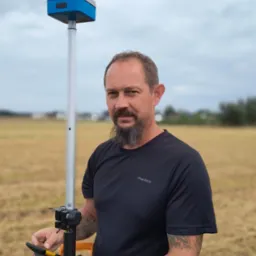 Mężczyzna z brodą, w czarnej koszulce, trzyma odbiornik GNSS na tyczce, na tle pola i pochmurnego nieba. Urządzenie pomiarowe.