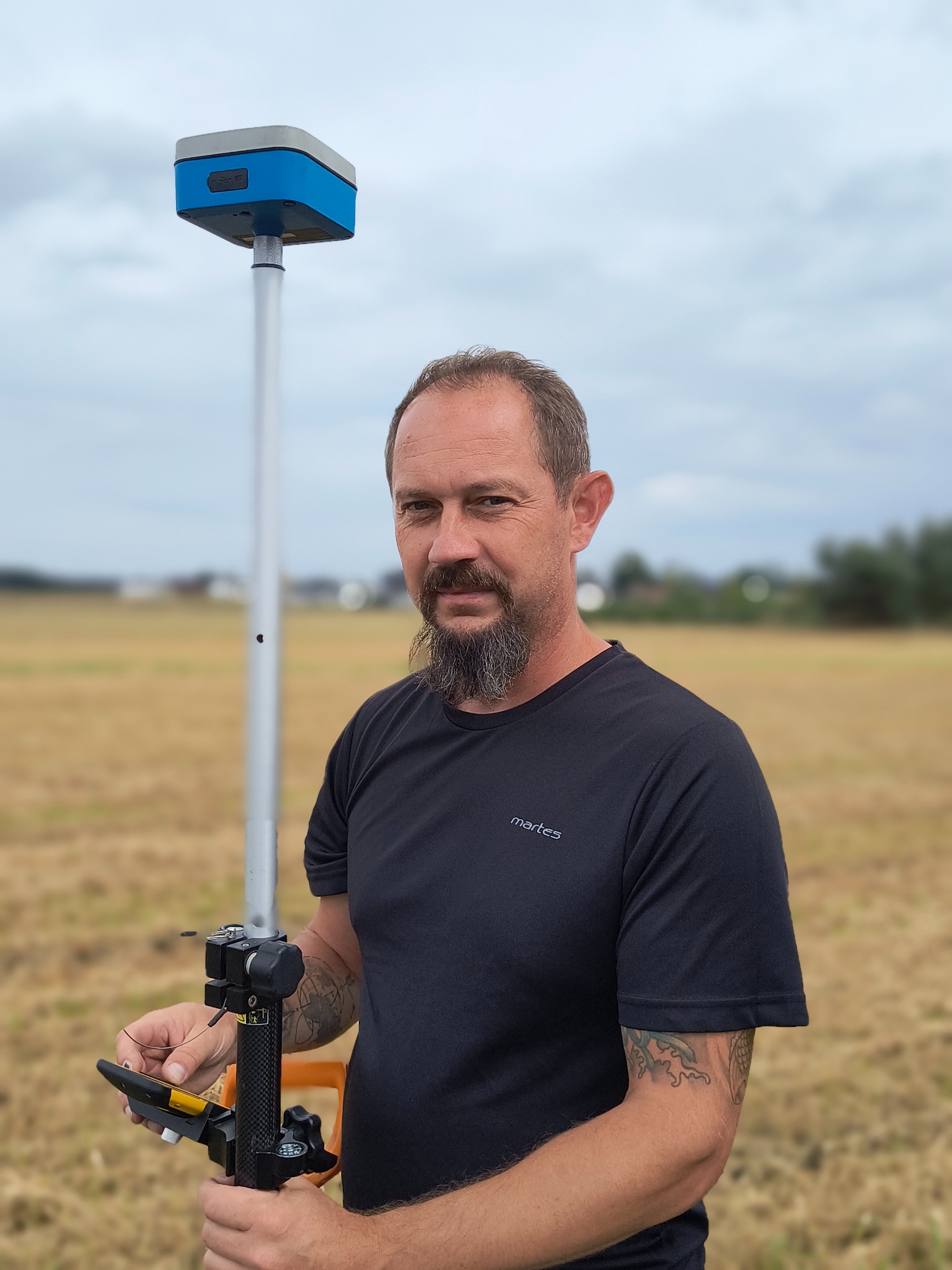 Mężczyzna z brodą, w czarnej koszulce, trzyma odbiornik GNSS na tyczce, na tle pola i pochmurnego nieba. Urządzenie pomiarowe.