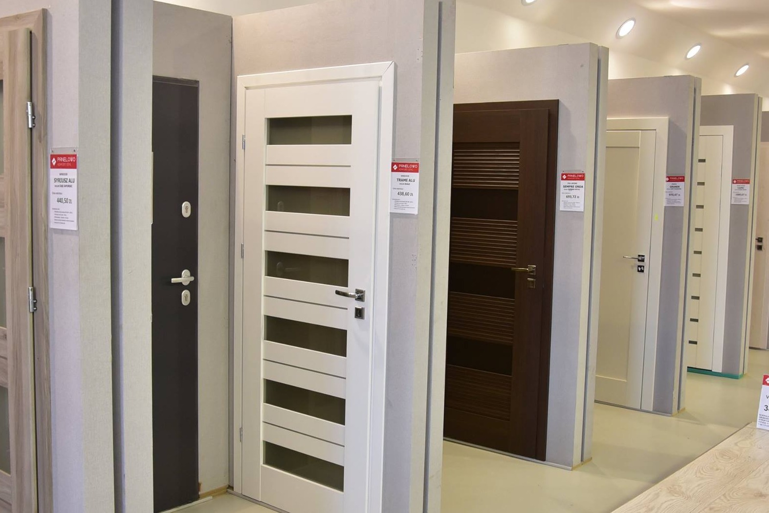 Ekspozycja różnorodnych modeli drzwi wewnętrznych w salonie. Widoczne drzwi białe z przeszkleniami, ciemne antywłamaniowe i inne warianty kolorystyczne.