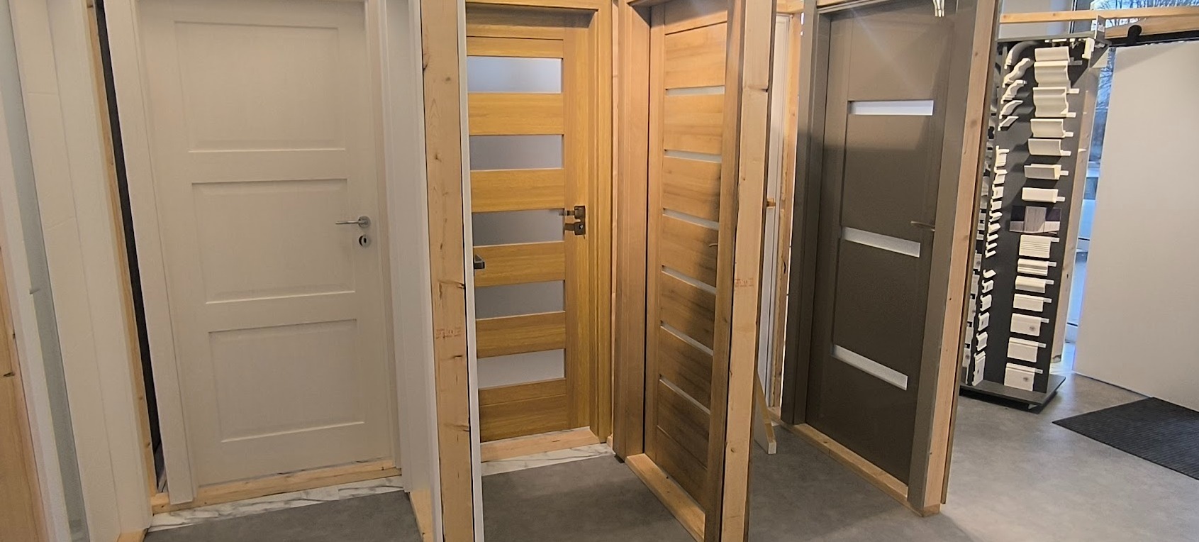 Ekspozycja trzech modeli drzwi wewnętrznych: białe, drewniane z przeszkleniami i szare. Widoczny fragment ekspozycji listew przypodłogowych.