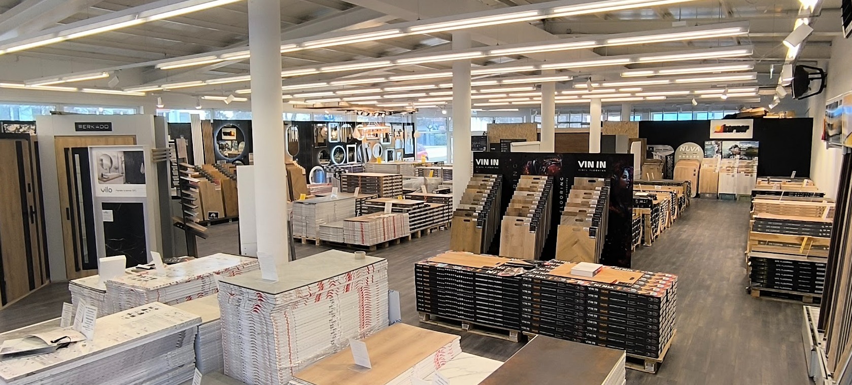 Szeroki kadr na ekspozycję paneli i drzwi w przestronnym salonie z jasnym oświetleniem. Widoczne wzorniki i opakowania produktów.