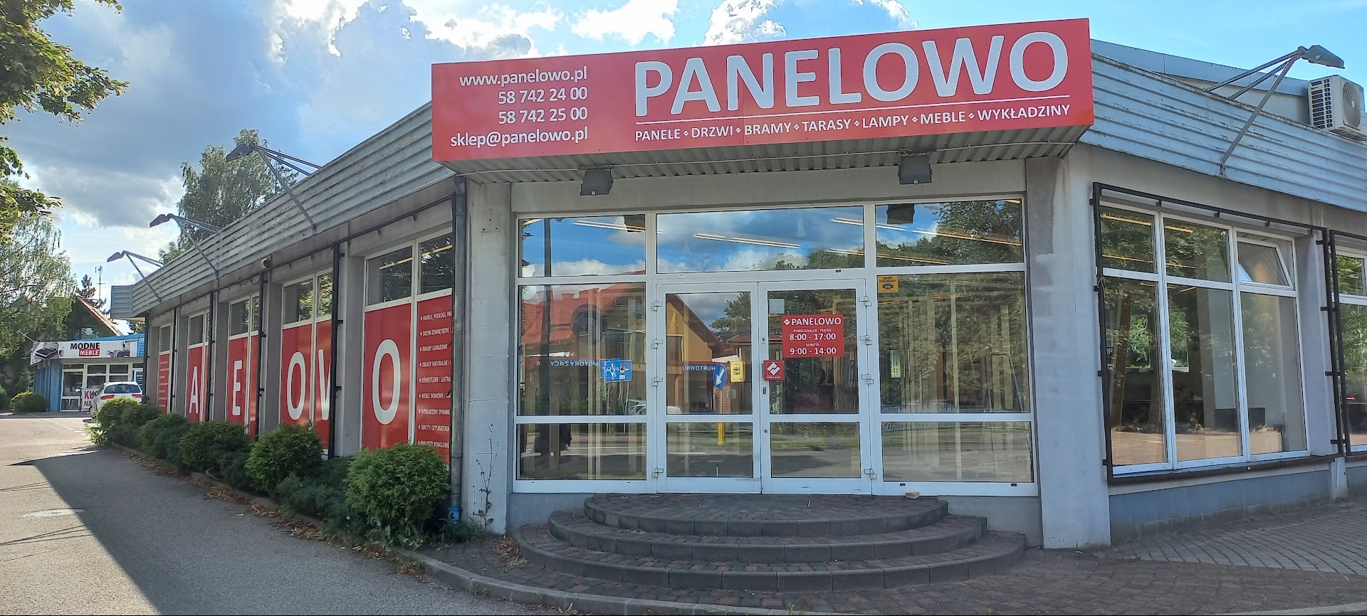 Fasada budynku firmy Panelowo z dużym szyldem reklamowym, prezentującym ofertę: panele, drzwi, bramy, tarasy, lampy, meble, wykładziny. Widoczne okna wystawowe i schody.