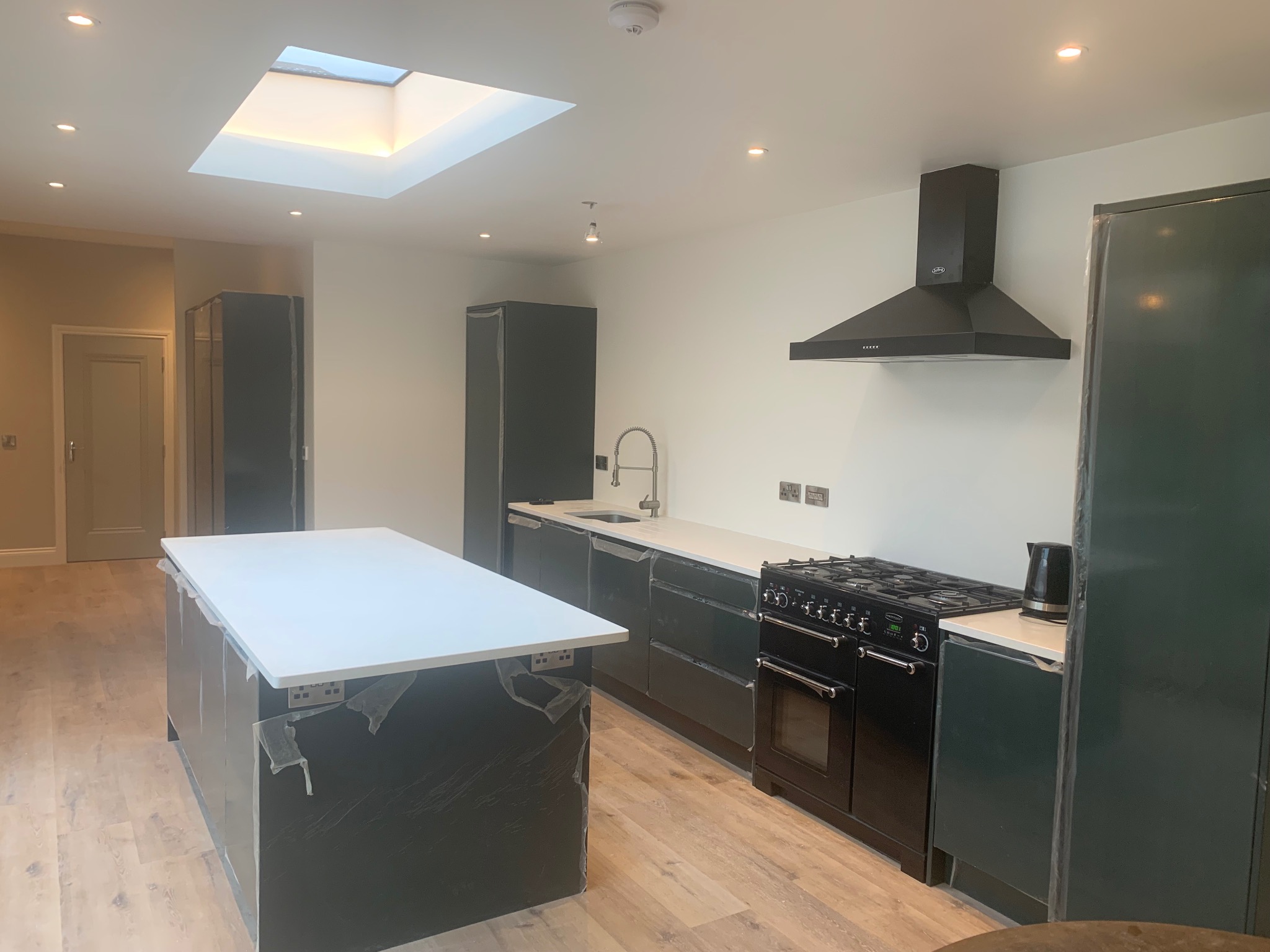 Nowoczesna kuchnia w trakcie wykończenia z wyspą, czarnymi szafkami i sprzętem AGD. Oświetlenie sufitowe i okno dachowe doświetlają wnętrze. Podłoga drewniana.