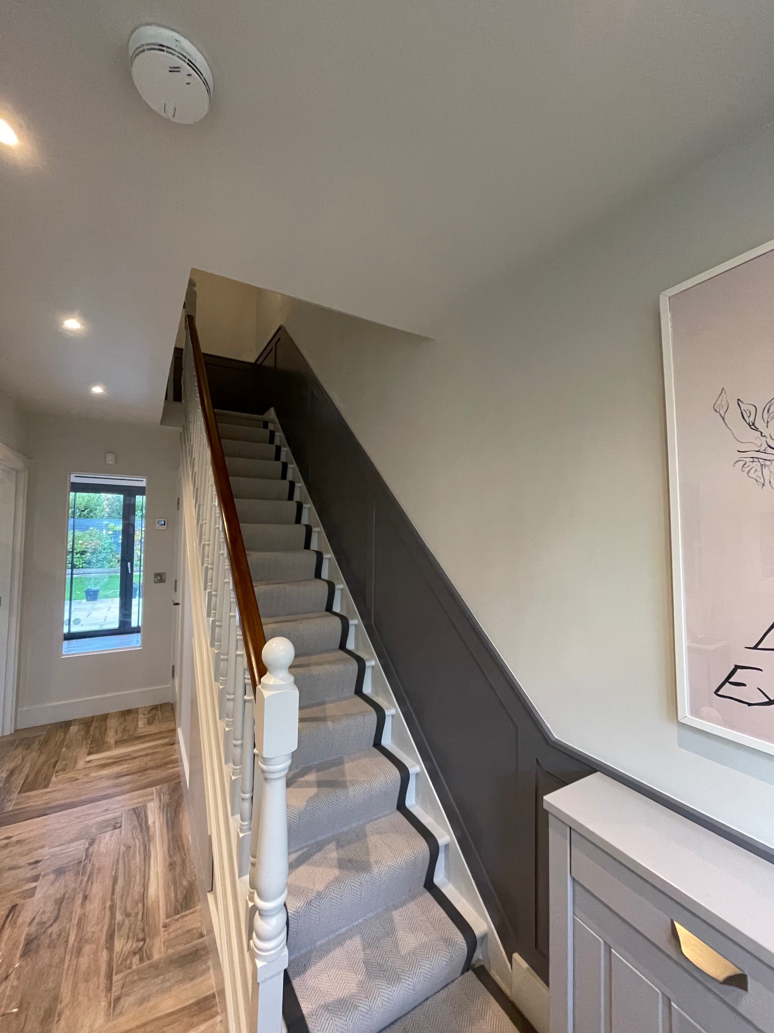 Klatka schodowa po renowacji: szare schody z czarnym obramowaniem, drewniana poręcz i szara boazeria. Na ścianie obraz i szafka. Ułożona podłoga.