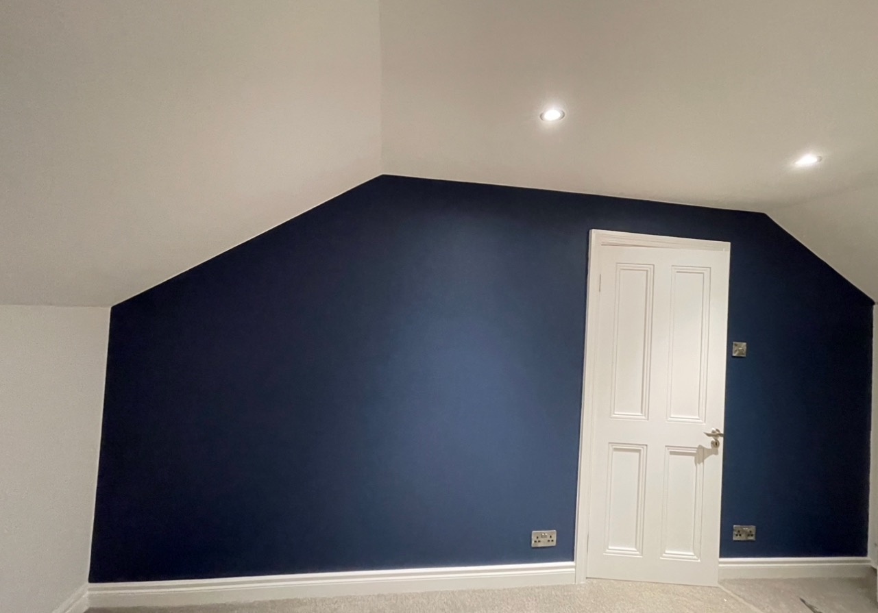 Pomalowane na granatowo poddasze ze skośnym sufitem i białą stolarką drzwiową. Widoczne gniazdka elektryczne i listwy przypodłogowe.