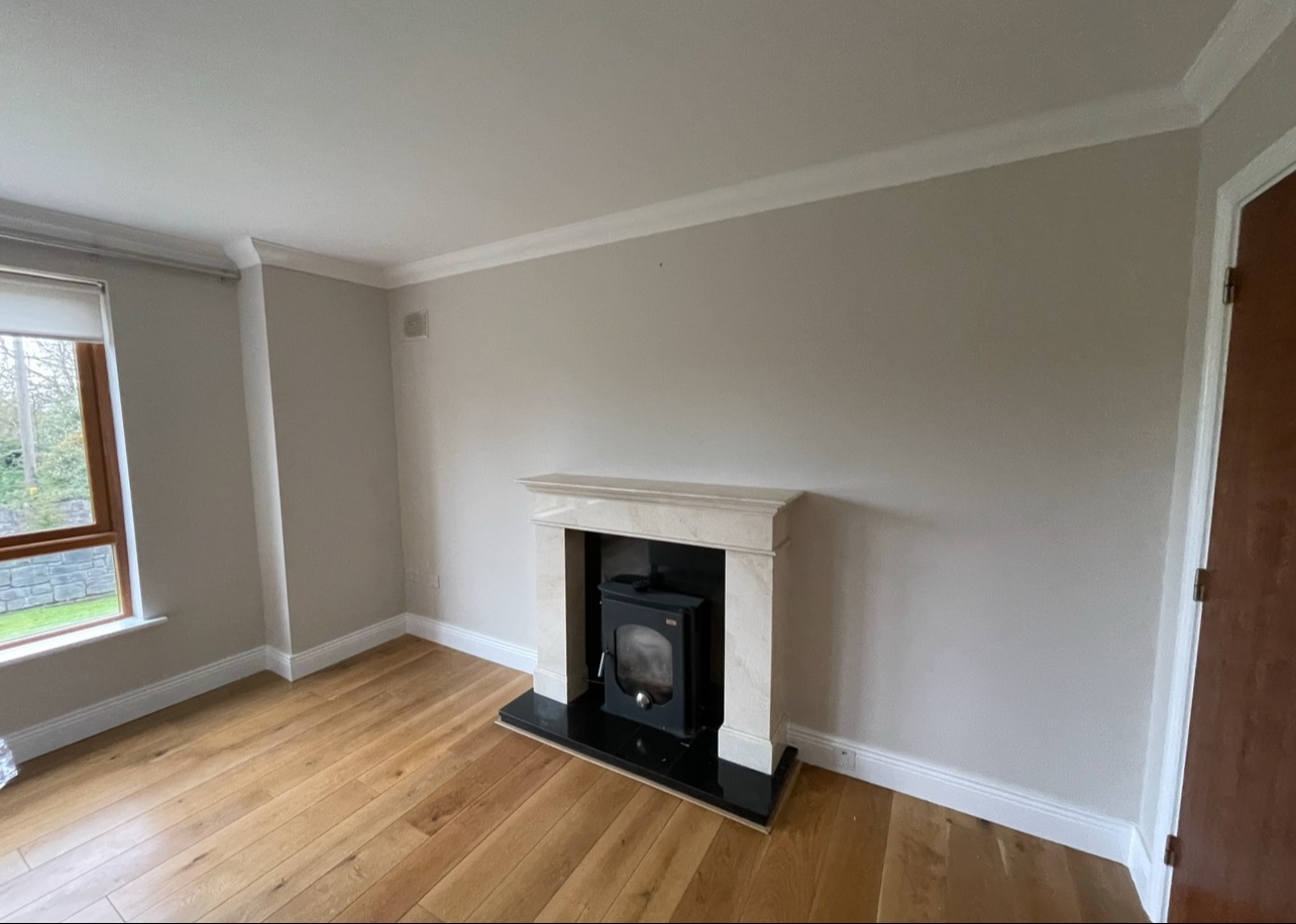 Jasny pokój po remoncie: drewniana podłoga, kremowe ściany z listwami, marmurowy kominek z czarnym wkładem. Okno po lewej stronie. Drzwi po prawej.