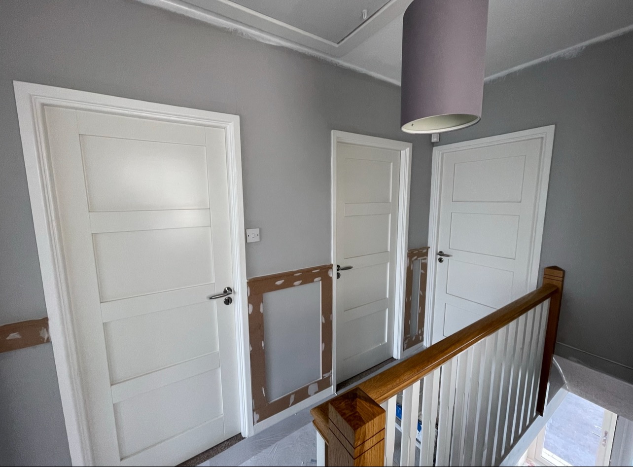 Klatka schodowa w trakcie remontu: szare ściany, białe drzwi z framugami w trakcie montażu, zabezpieczone ściany. Widok z góry na balustradę.