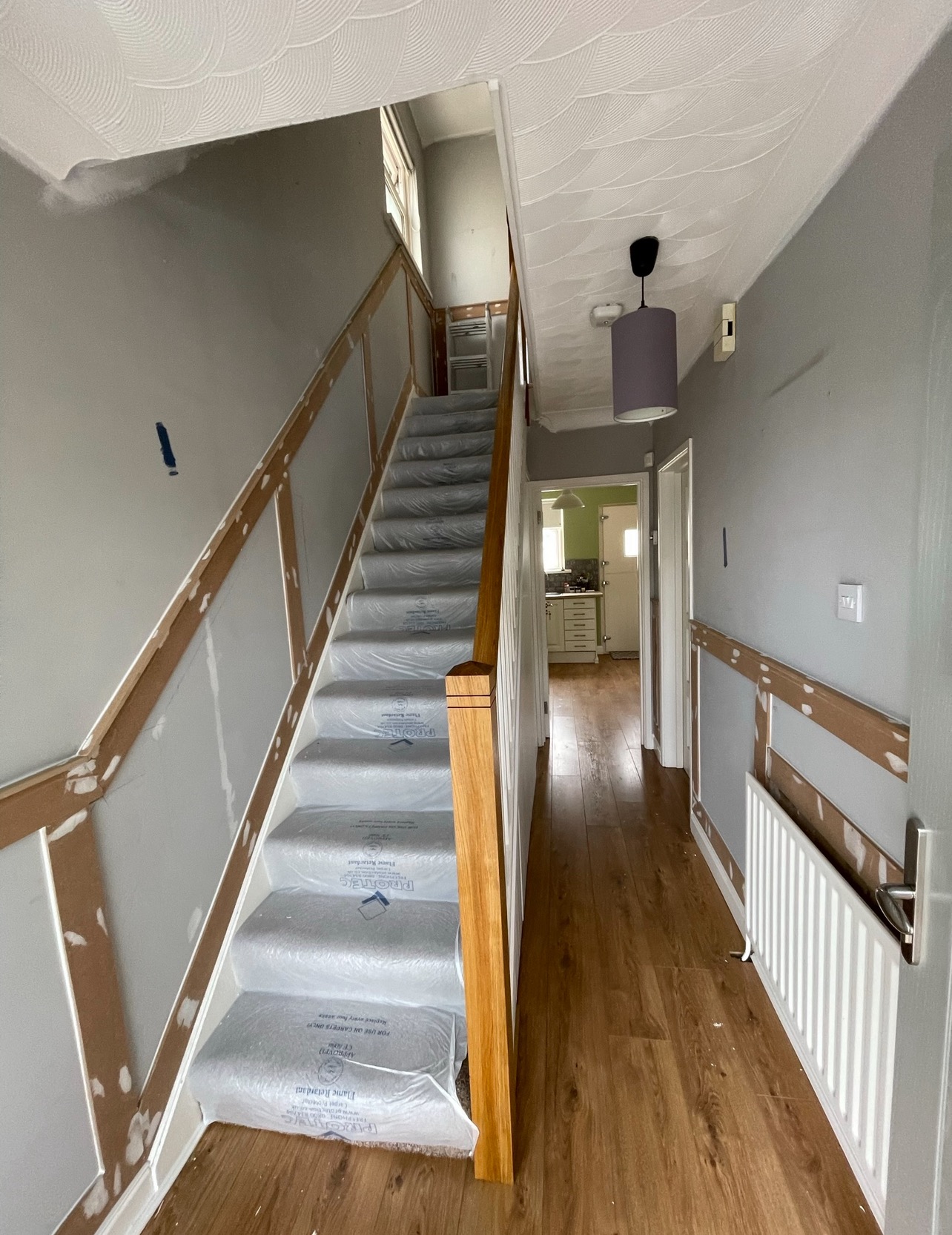 Korytarz w trakcie prac: zabezpieczone schody folią, ściany przygotowane do malowania, widoczne listwy przypodłogowe. Podłoga wyłożona panelami.