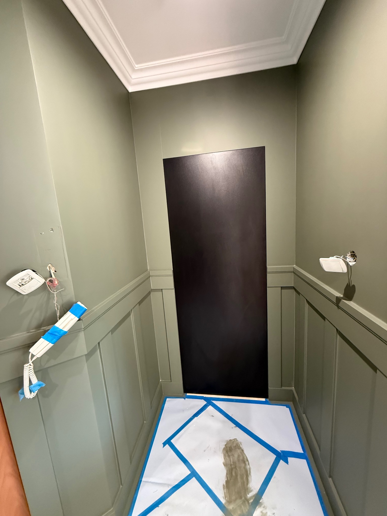 Ściana w odcieniach zieleni z dekoracyjną sztukaterią i zabezpieczoną podłogą. Widoczne przygotowania do prac wykończeniowych w pomieszczeniu.