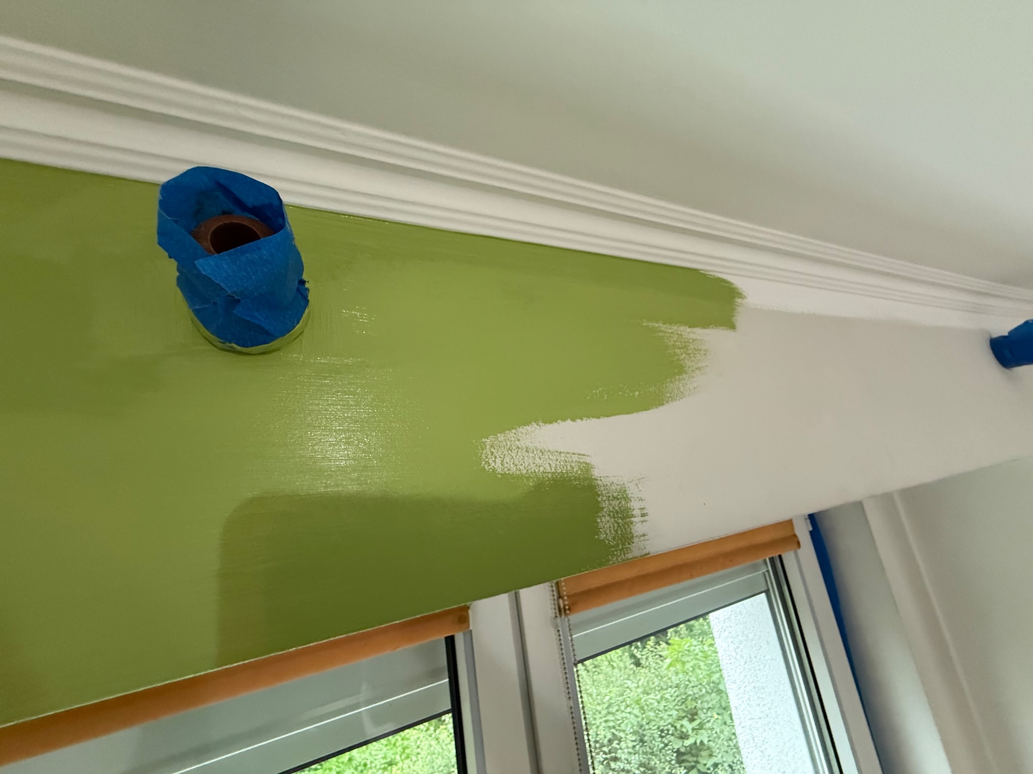 Malowanie ściany na zielono w trakcie. Część ściany pomalowana, druga biała. Gniazdko zabezpieczone taśmą malarską. Widoczny fragment okna.