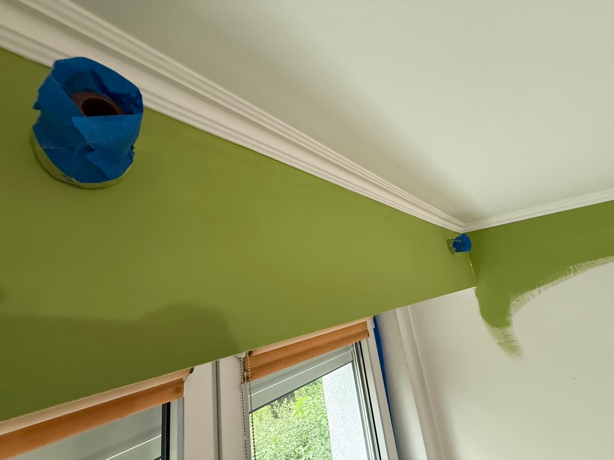 Fragment wnętrza w trakcie malowania: ściana w kolorze zielonym, biały sufit z dekoracyjną listwą. Otwór w ścianie zabezpieczony taśmą malarską.