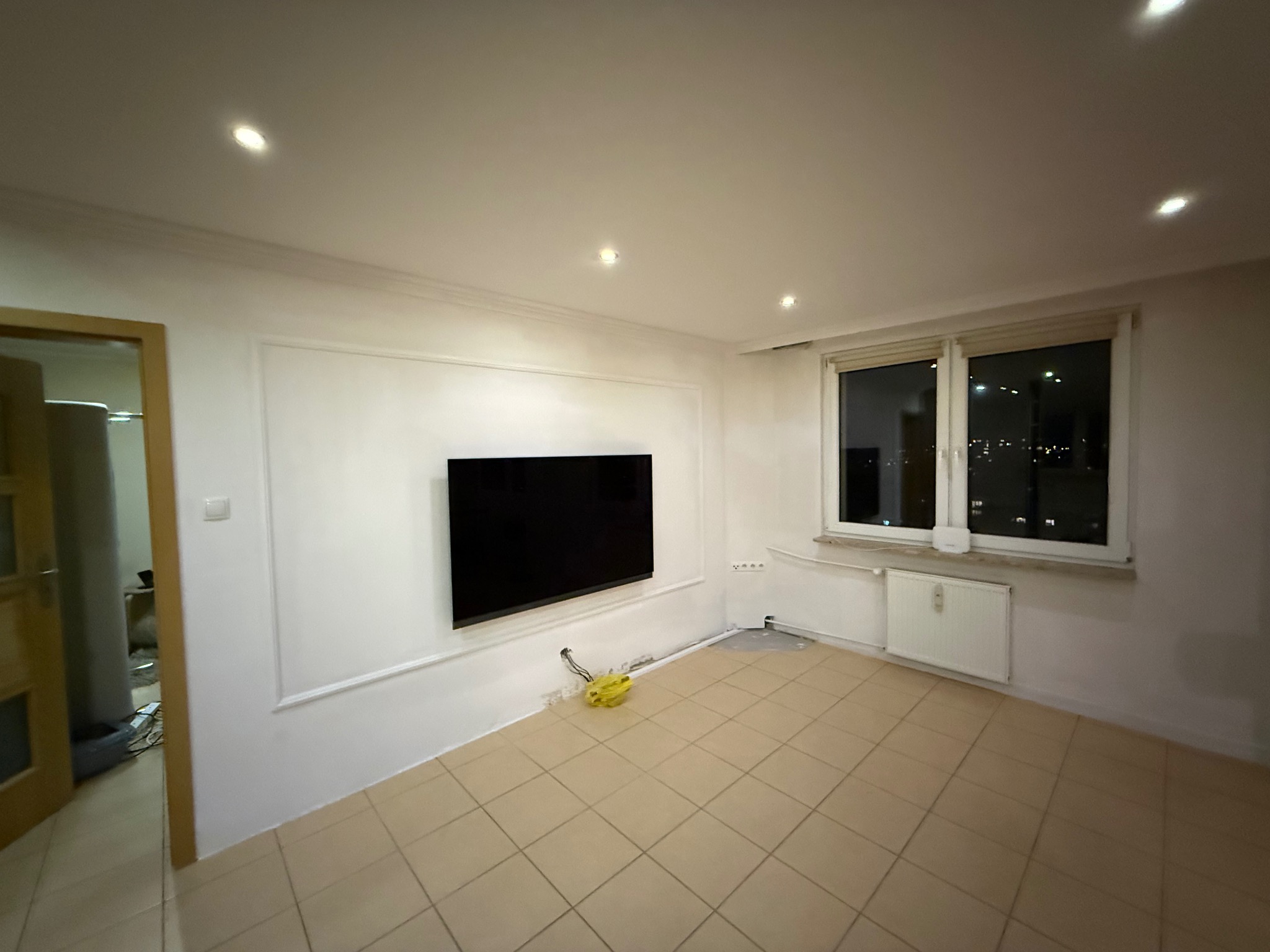 Pokój z beżową podłogą w kwadraty, białe ściany z dekoracyjną listwą, czarny telewizor na ścianie. Widoczne okno i grzejnik. Przygotowanie do prac wykończeniowych.