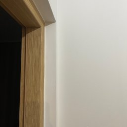 Lukasz Myszak - Gładka, biała ściana po malowaniu z widoczną ościeżnicą drzwiową z jasnego drewna. Fragment wykończonego wnętrza, minimalistyczny styl.